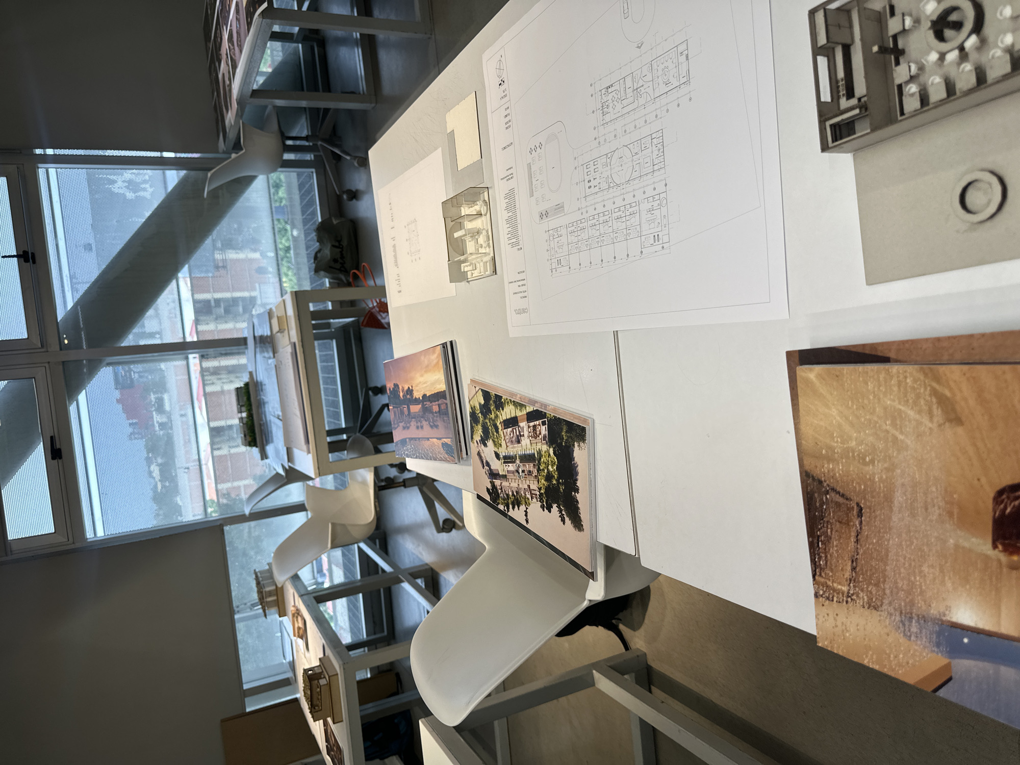 The image captures a bright, indoor space filled with architectural designs and models, likely an architecture studio. A large window on the left provides ample natural light and a view of the cityscape, including the distant buildings and clear sky, possibly a location in Mexico City, Mexico. The window is framed by a white metal structure.
The focus of the image is on a white table covered with architectural blueprints, photographs, and three-dimensional models. The blueprints display building layouts and technical drawings. Photographs showcase architectural projects or inspirations. Architectural models of varying sizes and materials are present.
Several white, modern chairs are positioned around the table, suggesting a collaborative workspace. The overall impression is of a creative environment dedicated to architectural design and presentation. The room appears to be well-organized and clean.