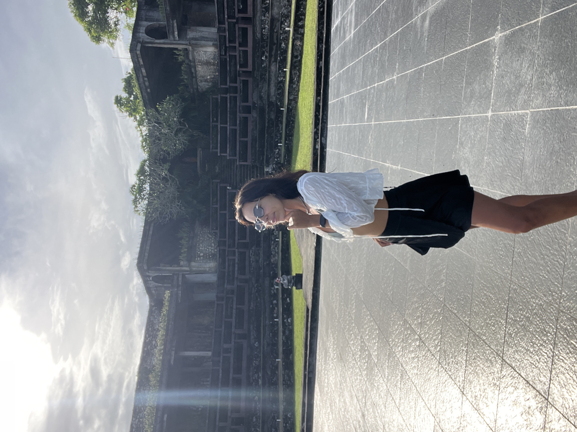 A young woman stands posed in a bright outdoor setting, facing slightly left of the camera. She has long dark hair, wears sunglasses, a white cropped long-sleeve shirt, and dark shorts. Her right hand is raised to her chin in a thoughtful or playful gesture, and she appears to be smiling or smirking subtly.
The background features an impressive, multi-tiered stone or concrete structure, possibly a temple or an ancient building complex, with lush green foliage and trees growing around and on it. This structure dominates the left and central background. In the foreground and midground, there is a large, light-colored paved area, possibly a courtyard or a shallow reflection pool, which shows considerable glare and reflections from the bright sun. Strips of vibrant green grass separate this paved area from the darker, textured stone walls of the main structure.
The scene is bathed in strong daylight, suggesting late morning or afternoon, with visible sun glare on the paved surface and some bright clouds in the sky, which occupies the upper left portion of the image. The architectural style, combined with the location context of Kuta Selatan, Indonesia, points to a Southeast Asian historical site or cultural landmark. There is no visible text in the image.