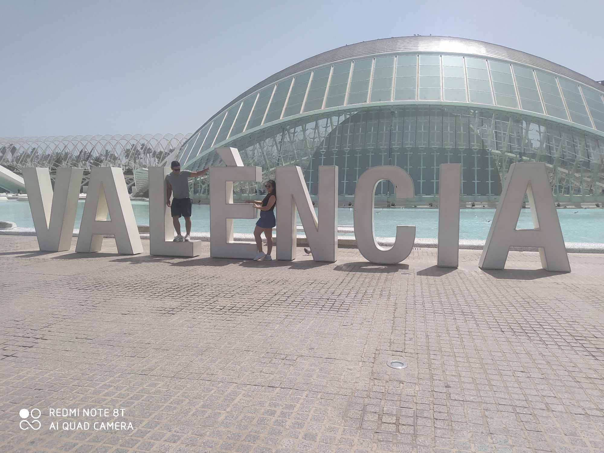 The image displays large white three-dimensional letters spelling "VALENCIA" on a paved surface. Two individuals are interacting with the letters. On the left, a man wearing a green t-shirt and dark shorts stands next to the letter "N" in "VALENCIA," with his left arm extended to touch it. On the right, a woman in a dark blue dress and white sneakers leans against the letter "N," her arms resting on it. Behind the letters, a large, modern building with a curved, glass facade is visible, reflecting the sky. The ground is made of patterned grey pavers, and in the distance, a body of water with white structures is present. The sky is a clear, light blue.