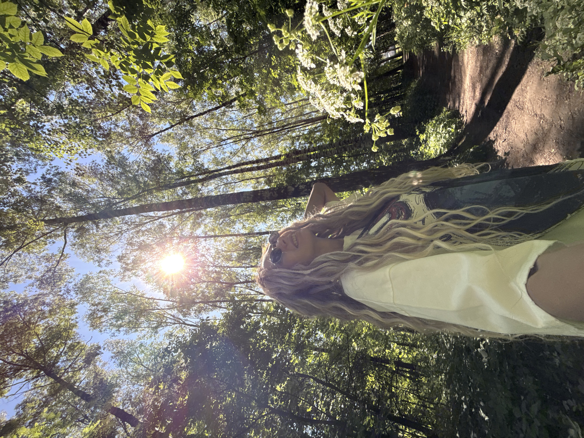 The image depicts a female subject standing within a sunlit wooded area in Rusanovo, Russia. The individual, characterized by long, wavy blonde hair and sunglasses, is positioned slightly off-center to the left, looking upward and to the right with a subtle smile. She is wearing a light-colored, long-sleeved garment, possibly an oversized shirt, with a darker design partially visible beneath her hair. Her left arm is raised, with her hand positioned near her head/ear area.
The environment is a forest or park dominated by tall, slender trees with green foliage, through which a bright blue sky is visible. A strong sunburst effect originates from the upper right quadrant of the image, indicating direct sunlight and creating lens flare. A narrow dirt path, partially shaded, winds into the background on the right side of the frame. Lush green undergrowth and vegetation line the path and forest floor. In the mid-ground on the right, a cluster of white flowering plants with umbrella-shaped inflorescences is prominently featured. Shadows from the trees are cast across the ground and path.