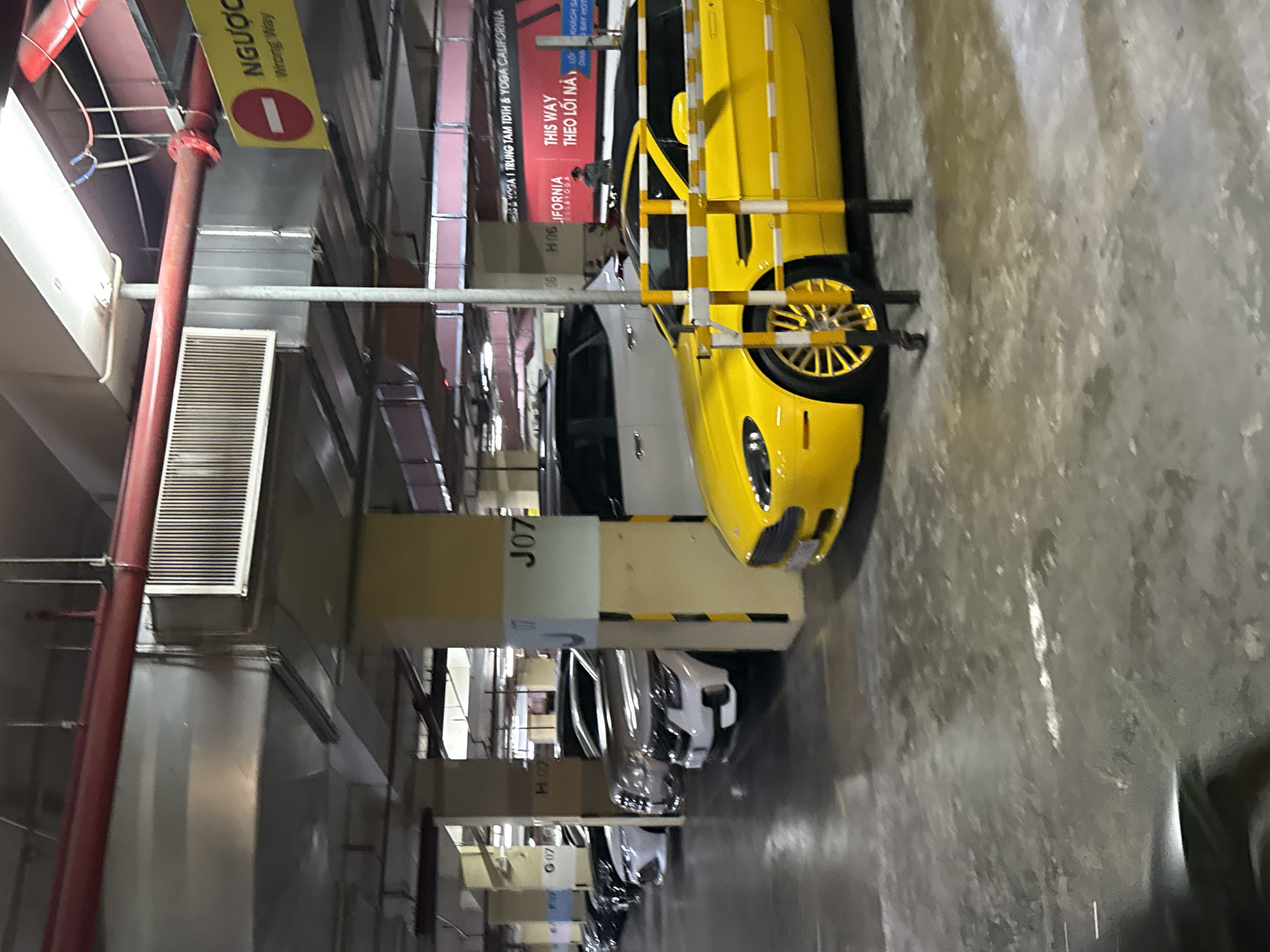 A bright yellow sports car is parked in an underground parking garage. The car has custom gold rims and a black roof. It is positioned in front of a row of other vehicles, including white SUVs and sedans. The parking garage features concrete pillars with overhead lighting and signage. One sign in Vietnamese reads "NGUOC WAY" with an English translation of "Wrong Way," indicating a traffic direction. Another sign is visible in the background with Vietnamese text and the words "YOGA CALIFORNIA." The floor of the garage is concrete and appears somewhat worn and wet in places, with reflective surfaces indicating the presence of water or oil. The overall lighting is dim, typical of an indoor parking structure. The scene is static, with no discernible action or interaction occurring.
