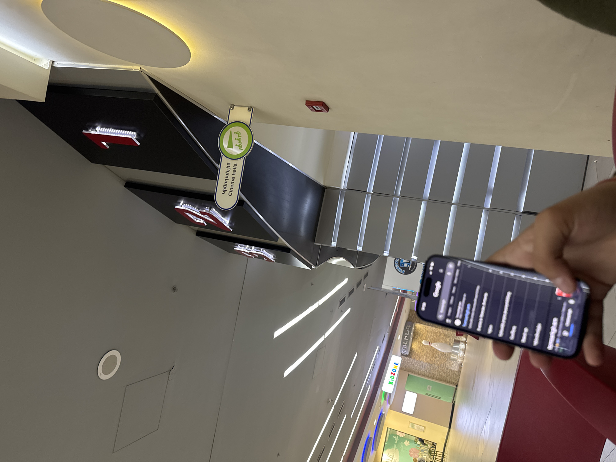 The image is taken inside a shopping mall, likely in Yerevan, Armenia, as indicated by the Armenian text on a sign that reads "Cinema halls" and on the phone screen. The mall's interior features a modern design with a gray ceiling, recessed lighting, and directional signage for "Cinema halls" illuminated with LED lights. The floors are light-colored, possibly tile or polished concrete, with linear ceiling lights running lengthwise. A set of stairs with gray railings and light-colored risers is visible in the background. In the foreground, a hand holds a smartphone. The screen displays a Google search interface with multiple tabs open. Visible in the mall environment are stores, including one labeled "KIDZONE," along with decorative elements like a bowling pin display. The mall appears modern and well-lit, suggesting a contemporary shopping experience. The image seems to be captured from a slightly elevated perspective, possibly from a mezzanine level or upper floor of the mall.