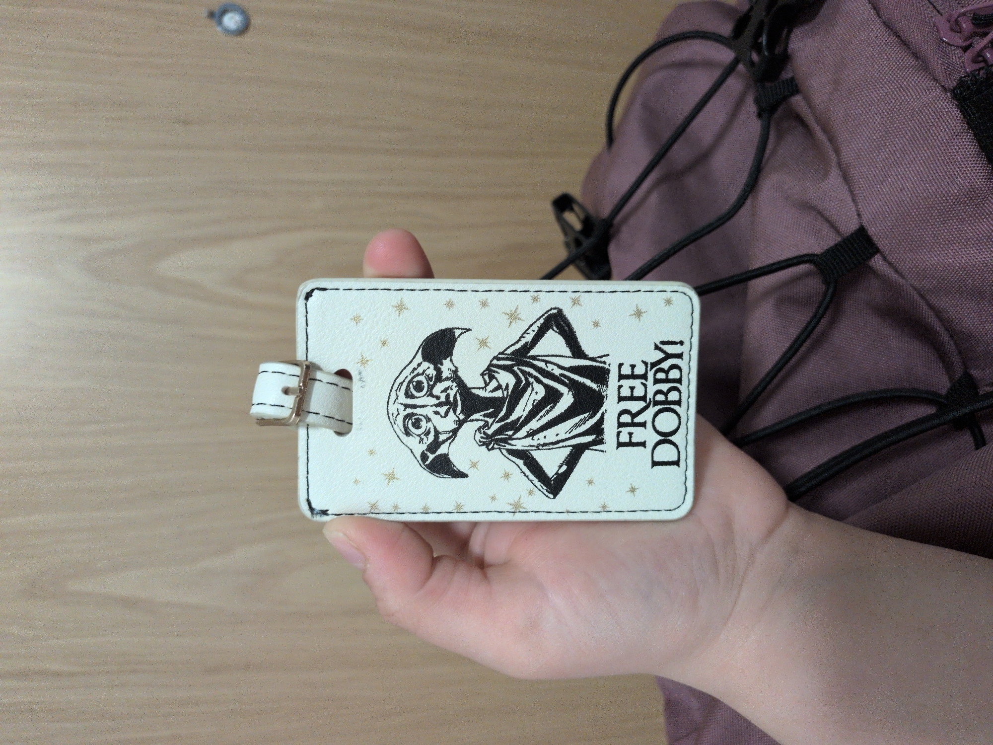 A pale-skinned hand holds a white, rectangular luggage tag featuring a black-and-white illustration of Dobby, the house-elf from Harry Potter. Dobby is depicted with large ears, wide eyes, and is wearing a simple cloth. Below the illustration, the text "FREE DOBBY!" is printed in black capital letters. Small, faint gold-colored starbursts are scattered across the tag's background. The tag has a black stitched border and a white strap with a gold-colored buckle emerging from the top. The top-left edge of the tag shows minor dark discoloration. The hand grasps the left side of the tag. In the upper background, a light brown wooden surface with vertical grain is visible. The lower background shows a crumpled, dusky purple fabric object, likely a bag or backpack, with black elastic cords woven into its surface and a partial view of a purple zipper pull. The image does not contain any visual information to determine its specific geographical location as Vantaa, Finland.