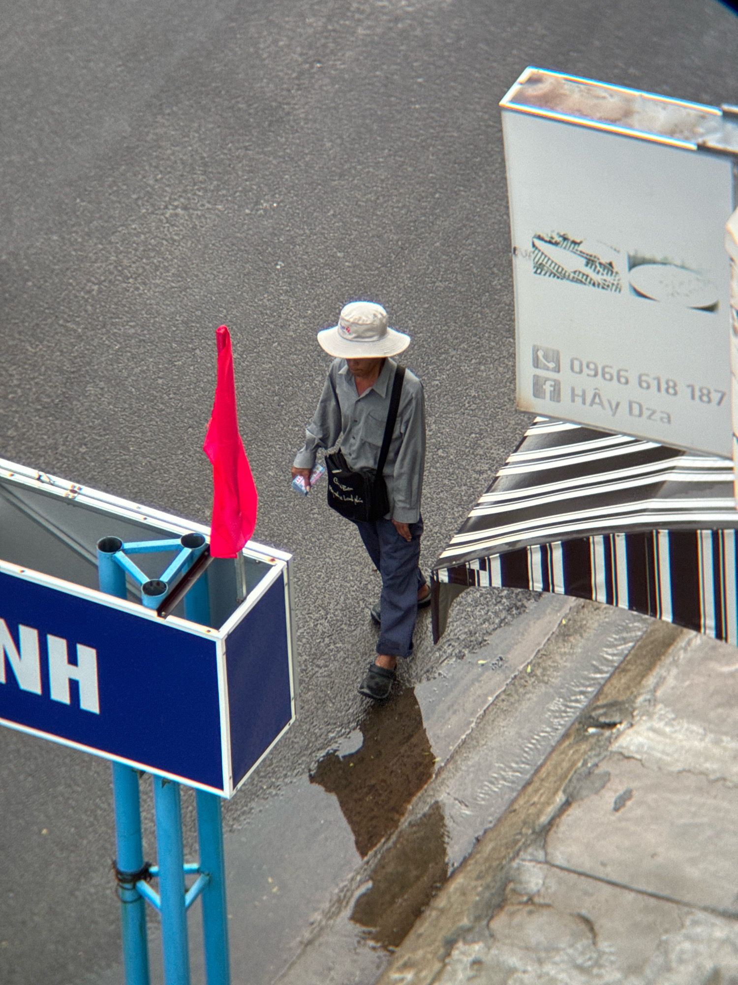 The image captures an overhead view of a street scene in Nha Trang, Vietnam. A person wearing a wide-brimmed hat, a gray shirt, and blue pants walks on a wet asphalt surface. The individual carries a dark bag slung across their body and holds a small, white object in their right hand. To the left, a blue sign with white lettering is partially visible. A bright red flag attached to a blue frame stands near the sign. In the background, a white sign with black text displays a phone number and what appears to be a business name. A striped awning is positioned to the right of the white sign. The ground is wet, suggesting recent rain, and the scene is illuminated by natural light.