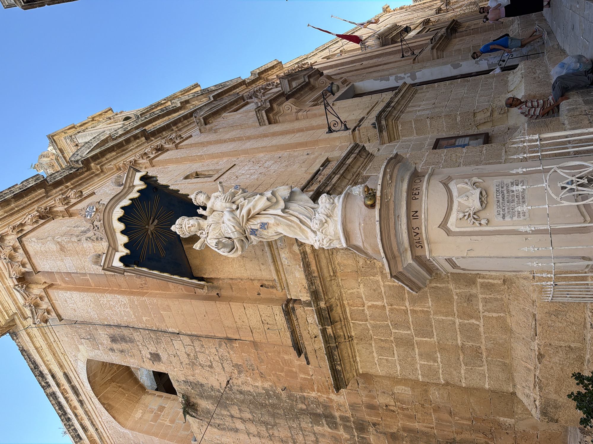 The image shows a statue of the Virgin Mary holding the infant Jesus, positioned on a pedestal outside a building. The statue is made of white stone and is intricately carved. The building appears to be a historic religious structure, with weathered stone walls and architectural detailing. A portion of a street or alleyway is visible, with a few people walking and sitting. The sky is clear and blue. The scene is set in the city of Mdina, Malta, likely in a narrow street. There is an inscription on the pedestal that reads "SALVS IN PERICULOS". The overall impression is one of historical and religious significance.