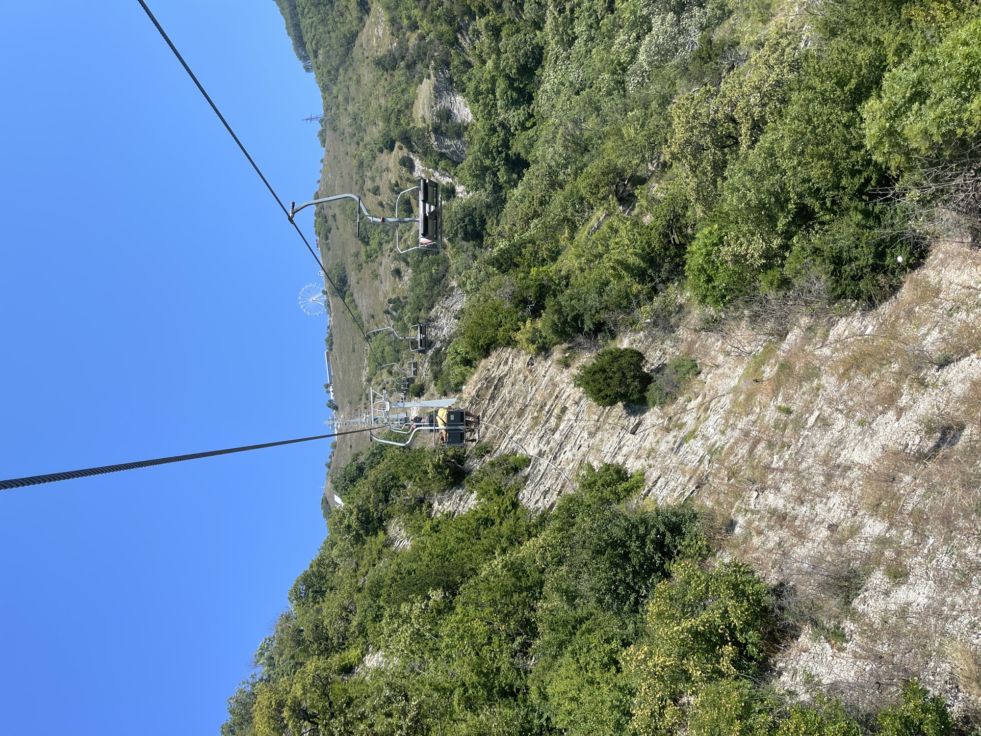 The image shows a cable car ascending a steep, rocky mountain covered in lush green vegetation. The cable car has multiple empty seats, with one visible car carrying two passengers wearing casual clothing. In the distance, atop the mountain, a Ferris wheel and some buildings are visible. The sky is clear and blue, indicating a sunny day. The scene is set in Gelendzhik, Russia, and depicts a popular tourist attraction offering panoramic views of the surrounding landscape. The mountain features visible stratification in the rock formations.