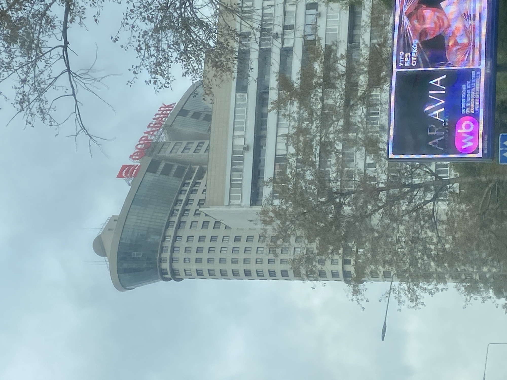 The image shows a wide shot of a modern, multi-story building in Moscow, Russia, under a cloudy sky. The building features a distinctive curved facade with numerous windows. A red neon sign that reads "ФОРА-БАНК" (FORA-BANK) is visible at the top of the building. In the foreground, a large digital billboard displays an advertisement. The billboard features the text "ARAVIA" and a woman's face with the text "УТРО БЕЗ ОТЕКОВ" (Morning without swelling). The billboard also includes the "wb" logo. Trees with sparse autumn foliage are visible in front of and around the building, partially obscuring the lower floors. Two streetlights are also present in the scene. The overall impression is an urban cityscape on an overcast day, with commercial advertising dominating the lower portion of the frame.