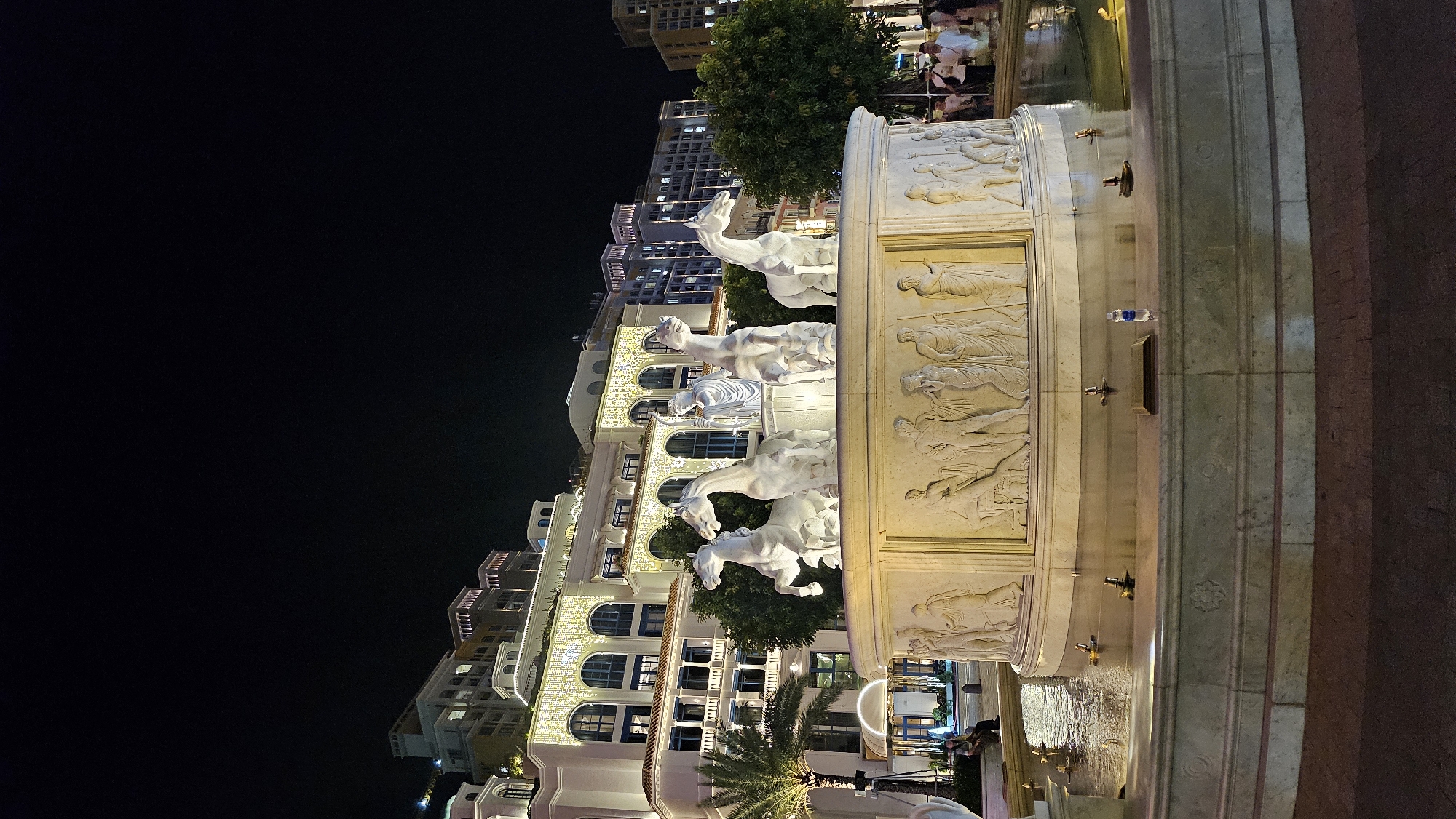 The image shows a large, ornate fountain at night in the city of Phú Quốc, Vietnam. The fountain is made of white marble and features sculptures of horses and a charioteer, as well as relief carvings depicting mythological scenes. The fountain is surrounded by a pool of water, and in the background, there are several multi-story buildings illuminated by warm lighting. Some people are visible in the distance, walking and sitting near the fountain. The sky above is dark and clear.