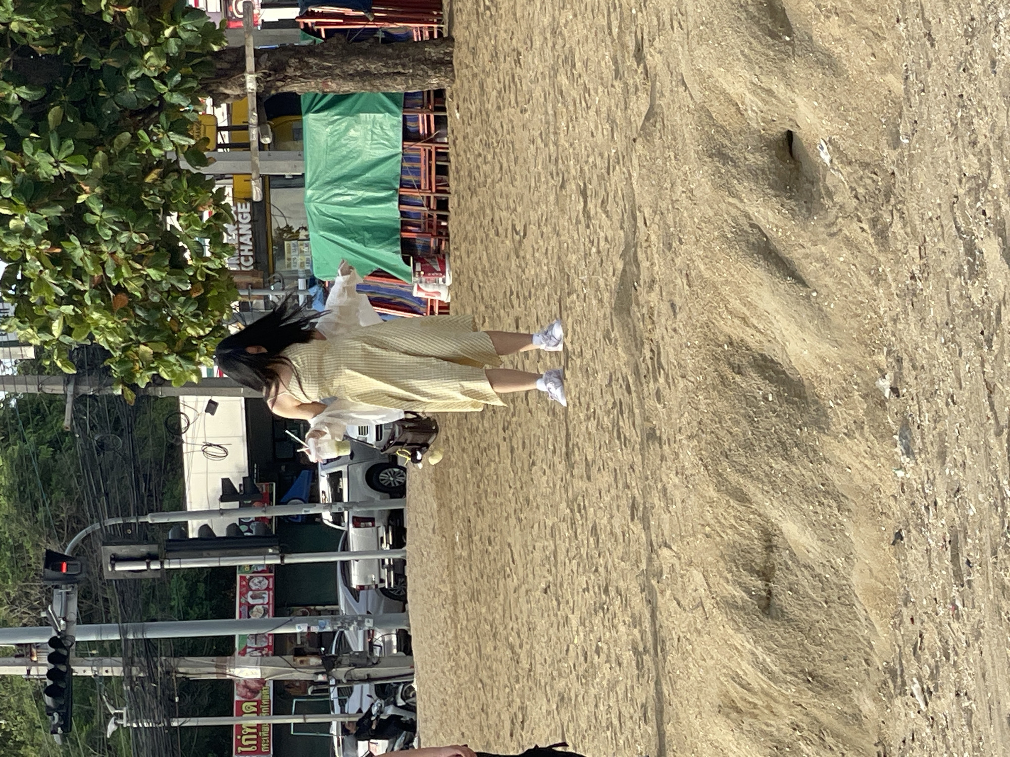 In Pattaya City, Thailand, on a sandy beach, a young woman stands in a light yellow checkered dress and a white open cardigan. She holds a clear plastic cup with a straw in her right hand. She is wearing white sneakers. Her dark hair is flowing. In the background, there are traffic lights and overhead wires, indicating a street close to the beach. A green tarp covers a set of chairs. The ground is covered in sand with some debris.