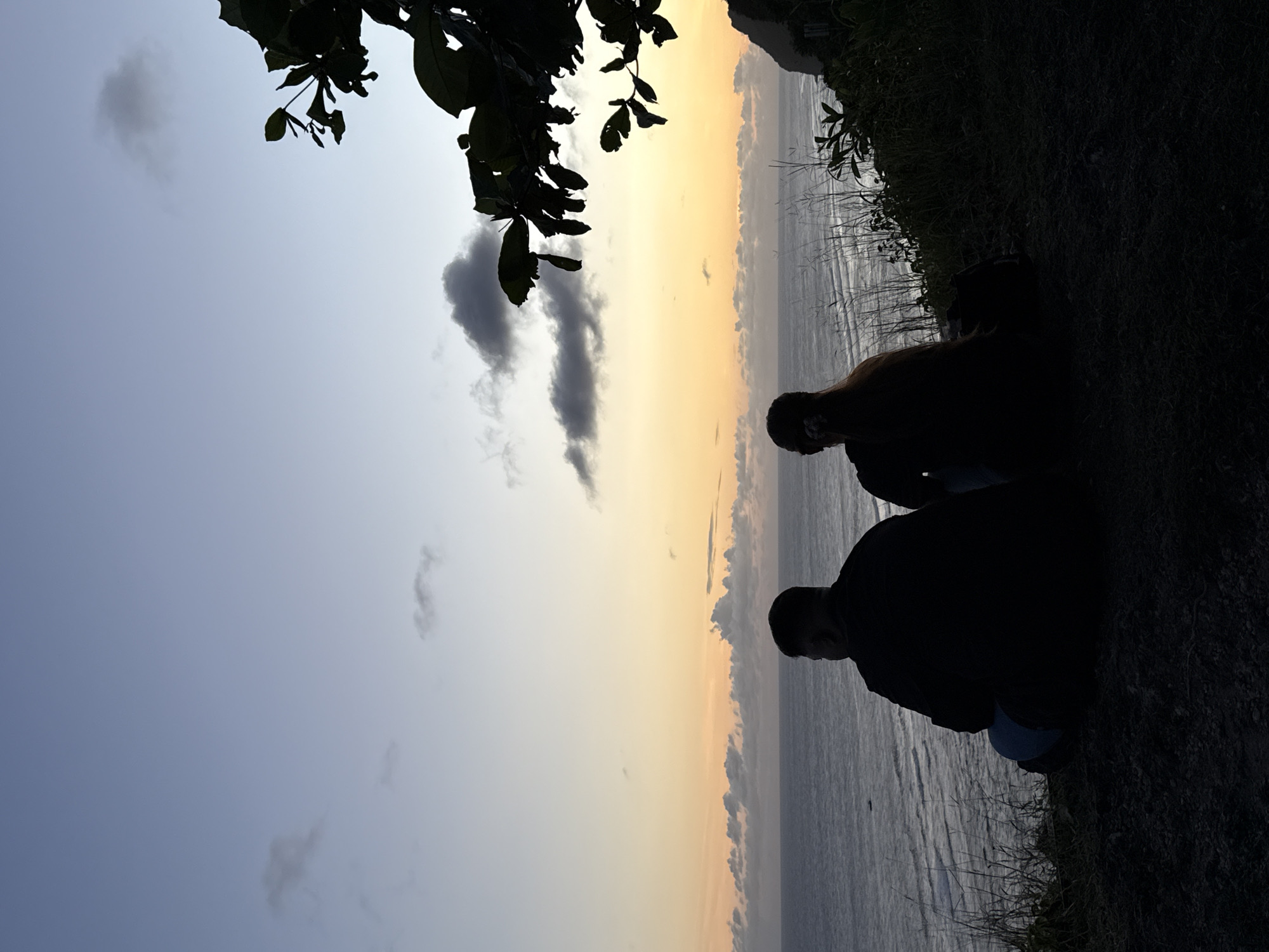 The image captures a landscape at sunset, likely in Ungasan, Indonesia, given the coastal environment. Two figures are silhouetted against the sky and sea, appearing to be crouching or sitting near the edge of a cliff or embankment overgrown with vegetation. The sky transitions from a light blue to a warm orange-yellow hue near the horizon. The sea below reflects the colors of the sky. There are some small clouds in the sky. Some tree branches and leaves are visible in the upper left corner of the image. The foreground is dark, highlighting the silhouettes of the figures and the grassy edge of the cliff. The photo has been rotated horizontally.