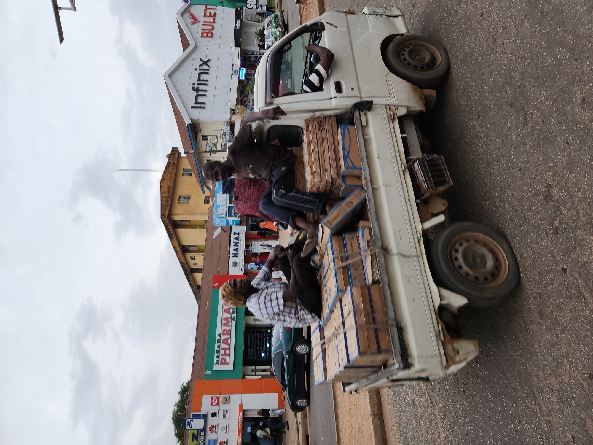 A white mini-truck drives on a road in Jos, Nigeria, carrying several people in its open bed, among stacks of packaged items. The truck, with visible wear and rust, has a person visible in the driver's seat. The road appears to be paved. Buildings line the street behind the truck, including a NASARA PHARMACY, HAMAZ store, and an Infinix store with a "BULET" sign. The sky is overcast.
