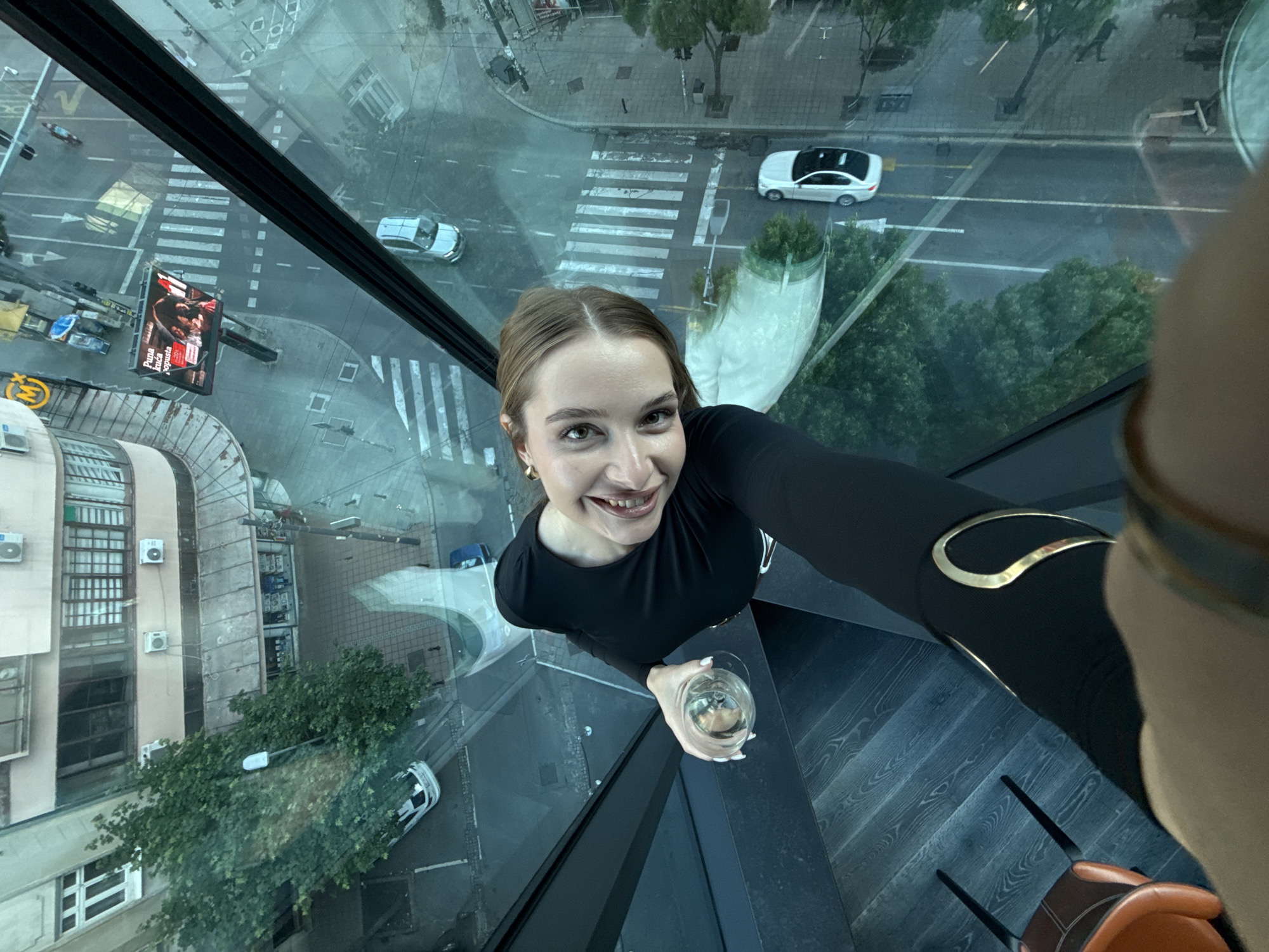 A high-angle selfie shows a young woman indoors, holding a glass of white wine, with a city view visible through the glass walls of the building. The location appears to be in Vracar Urban Municipality, Serbia, as suggested by the cityscape below.
The woman has light brown hair and is wearing a black top with gold embellishments on the sleeve. She is smiling and looking directly at the camera. In her left hand, she holds a glass containing a light-colored liquid, presumably wine.
Through the glass walls, there is an urban scene with buildings, streets, cars, and trees. Pedestrian crosswalks are visible on the streets below. A large advertising billboard is seen near a street intersection. The reflection of the city is visible on the glass walls around the woman. There is a dark wood floor and modern furniture in the room.