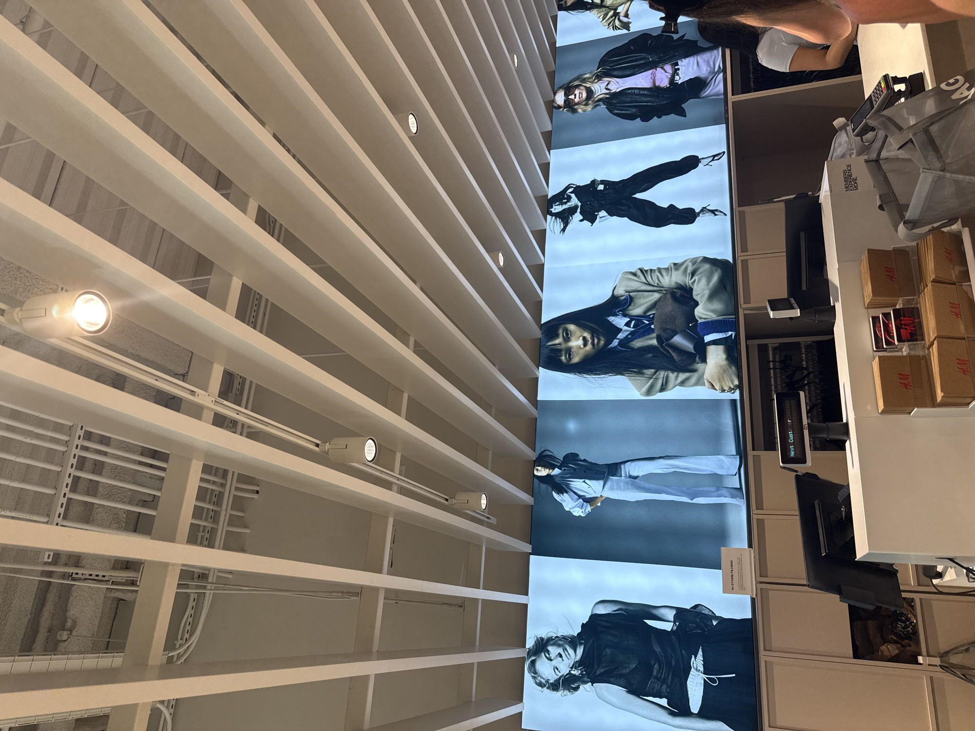 The image shows a retail store interior, likely H&M, with large digital advertisements featuring models. The advertisements are displayed on a wide screen across the upper portion of the image. Below the screen are checkout counters with registers, payment terminals, and retail bags. There are also stacks of gift cards and cardboard boxes with the H&M logo. In the foreground, part of a person's shoulder and head are visible, suggesting they are a customer. The overall environment is clean and well-lit, indicative of a modern retail space. The location is in Miami, United States of America.