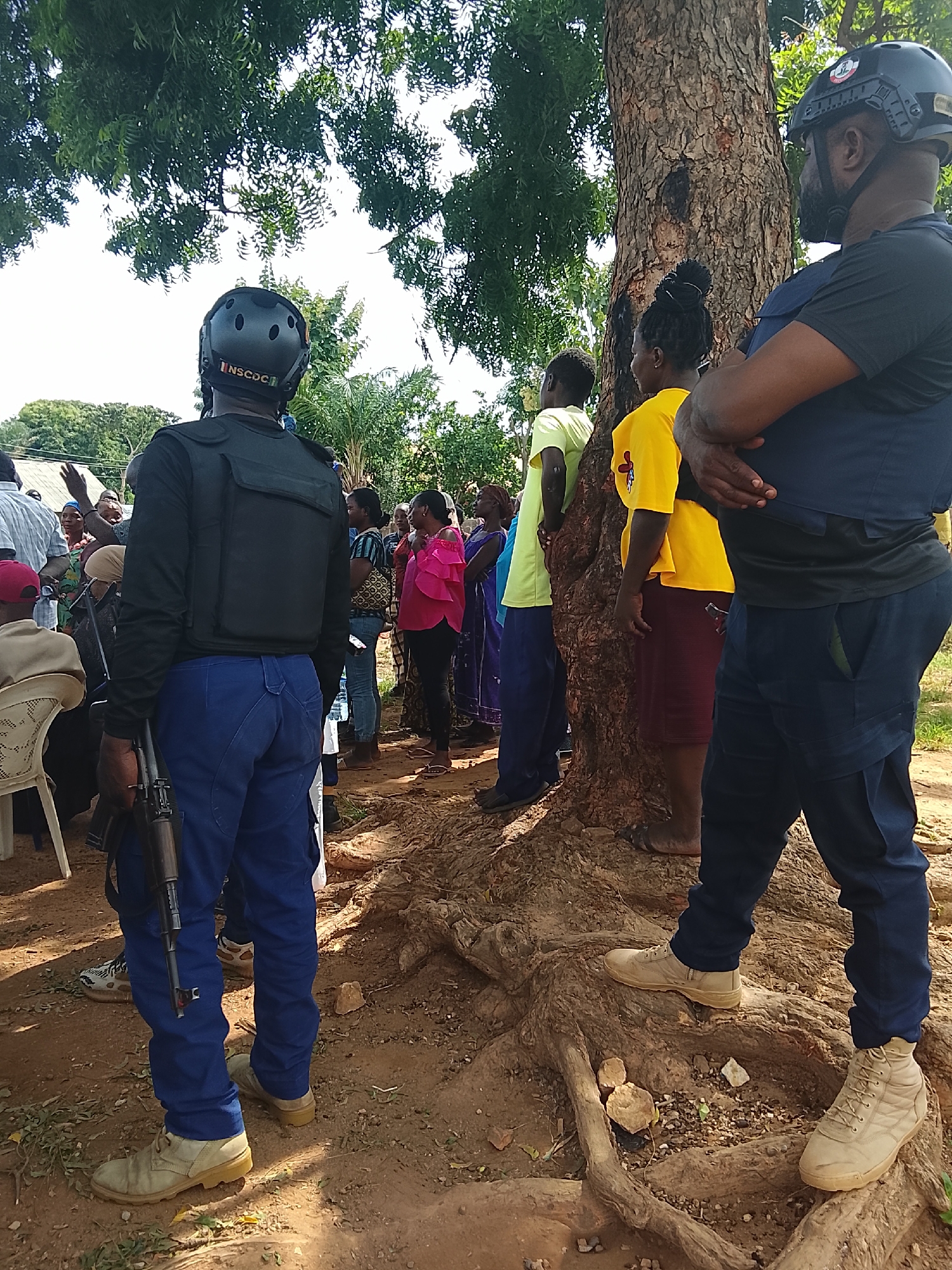 In an outdoor scene in Unknown, Unknown, a group of people are gathered around a large tree. A man wearing a black tactical vest, a black helmet with the inscription "NSCDC", and blue pants stands with his back to the camera, holding a rifle. To his right, another man in a dark blue shirt and dark pants stands with his arms crossed. Several other individuals, including women and children, are visible in the background, some appearing to be observing the situation. The ground is dirt and covered with tree roots and dry leaves. The environment suggests a rural or semi-rural setting with trees and greenery.