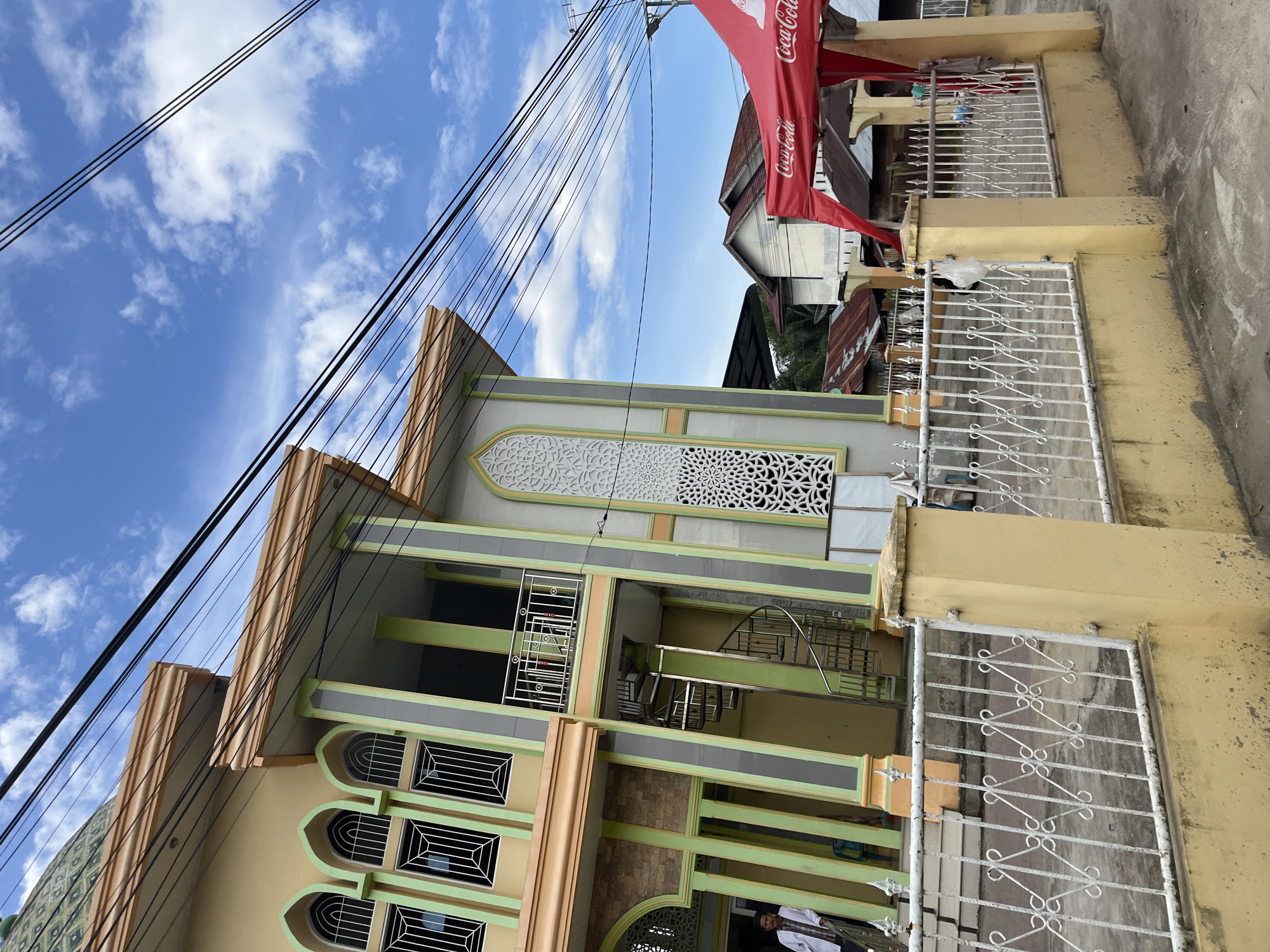 The image shows a mosque with a distinctive yellow and green facade, a green tiled dome, and decorative white window grilles. A spiral staircase is visible on the side of the building. In the foreground, a yellow concrete wall with a white metal fence runs along the street. A red tent with "Coca-Cola" branding is set up to the right. Several power lines are strung across the blue sky, which is dotted with white clouds. A man in a white shirt stands near the entrance of the mosque. The scene appears to be located in a somewhat urban environment, possibly in the city of Langsa, Indonesia, given the architectural style and the presence of a mosque.