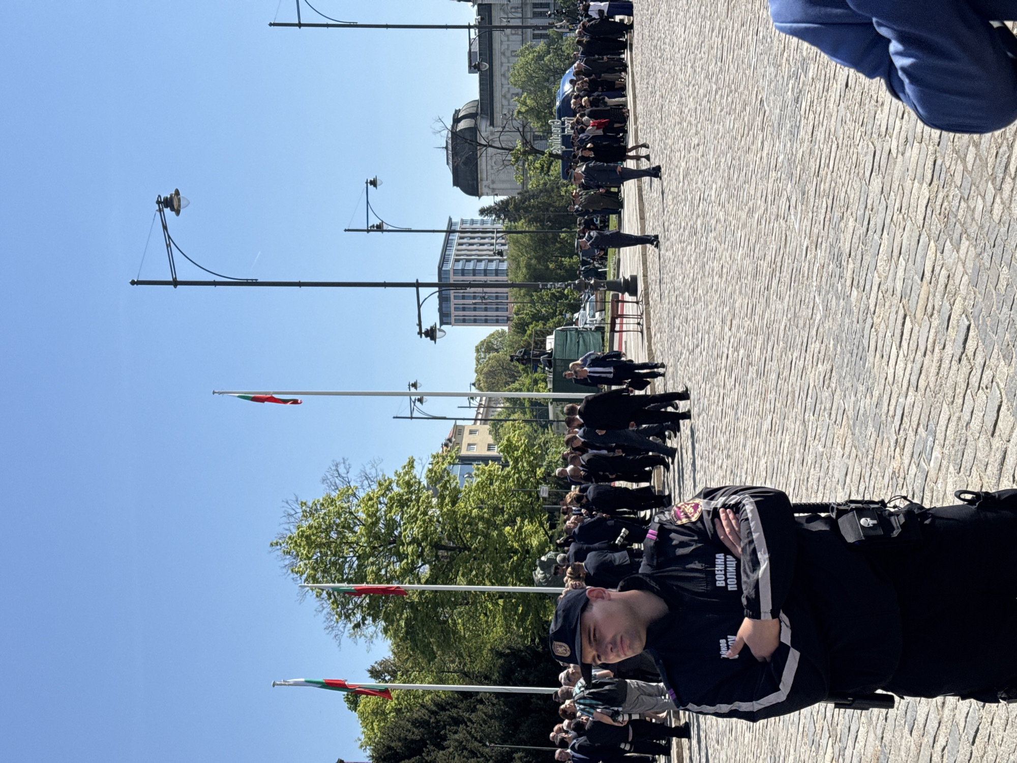 The image shows a scene in Sofia, Bulgaria. A cobblestone square is populated by a crowd of people, most of whom are dressed in dark suits. Several Bulgarian flags on flagpoles are visible. Lampposts line the square. In the foreground is a military police officer in a dark uniform with the inscription "ВОЕННА ПОЛИЦИЯ" (Military Police) on the sleeve, standing with arms crossed. In the background is a building with a dome.