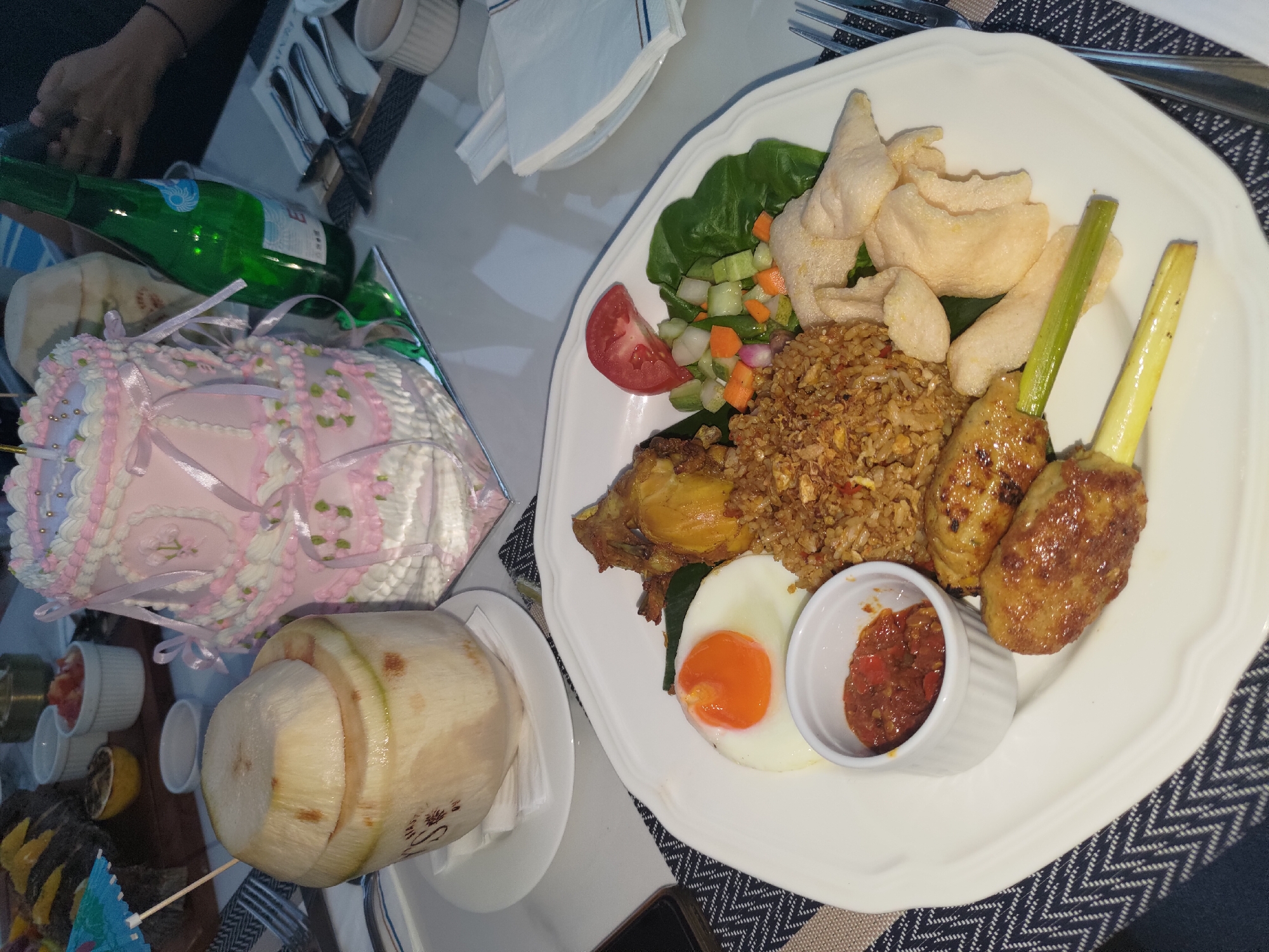 The image depicts a close-up, eye-level shot of a white plate filled with a meal, set on a table in what appears to be a restaurant in Kosambi Barat, Indonesia. The meal consists of fried rice, a fried egg with a visible orange yolk, satay skewers, fried chicken, a small dish of red chili paste, and a side salad with lettuce, chopped carrots, cucumber, and tomato. Several pieces of light-colored crackers are also present on the plate. In the background, to the left, is a whole coconut with its top cut off, serving as a beverage container, garnished with a small blue and white umbrella. Adjacent to the coconut is a tall, tiered pink cake decorated with white frosting, pink ribbon accents, and small green floral designs. A green glass bottle of sparkling water and cutlery are also visible on the table. The overall setting suggests a celebratory or dining occasion.
