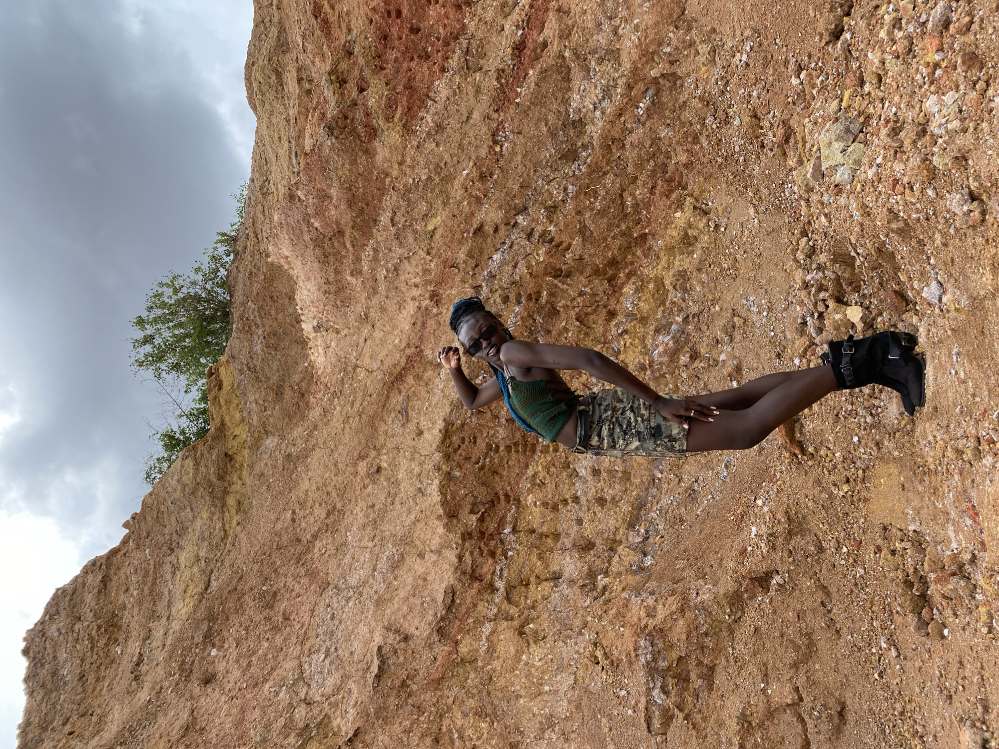 A young Black woman with braided hair and sunglasses poses playfully in front of a large, orange-brown rock formation under a cloudy sky. She is wearing a green, textured tank top and a camouflage skirt, with black boots. The terrain around her is rocky and uneven. The background appears to be a quarry or a similar geological site. The location is identified as Bar Jirgi Summa, Nigeria.