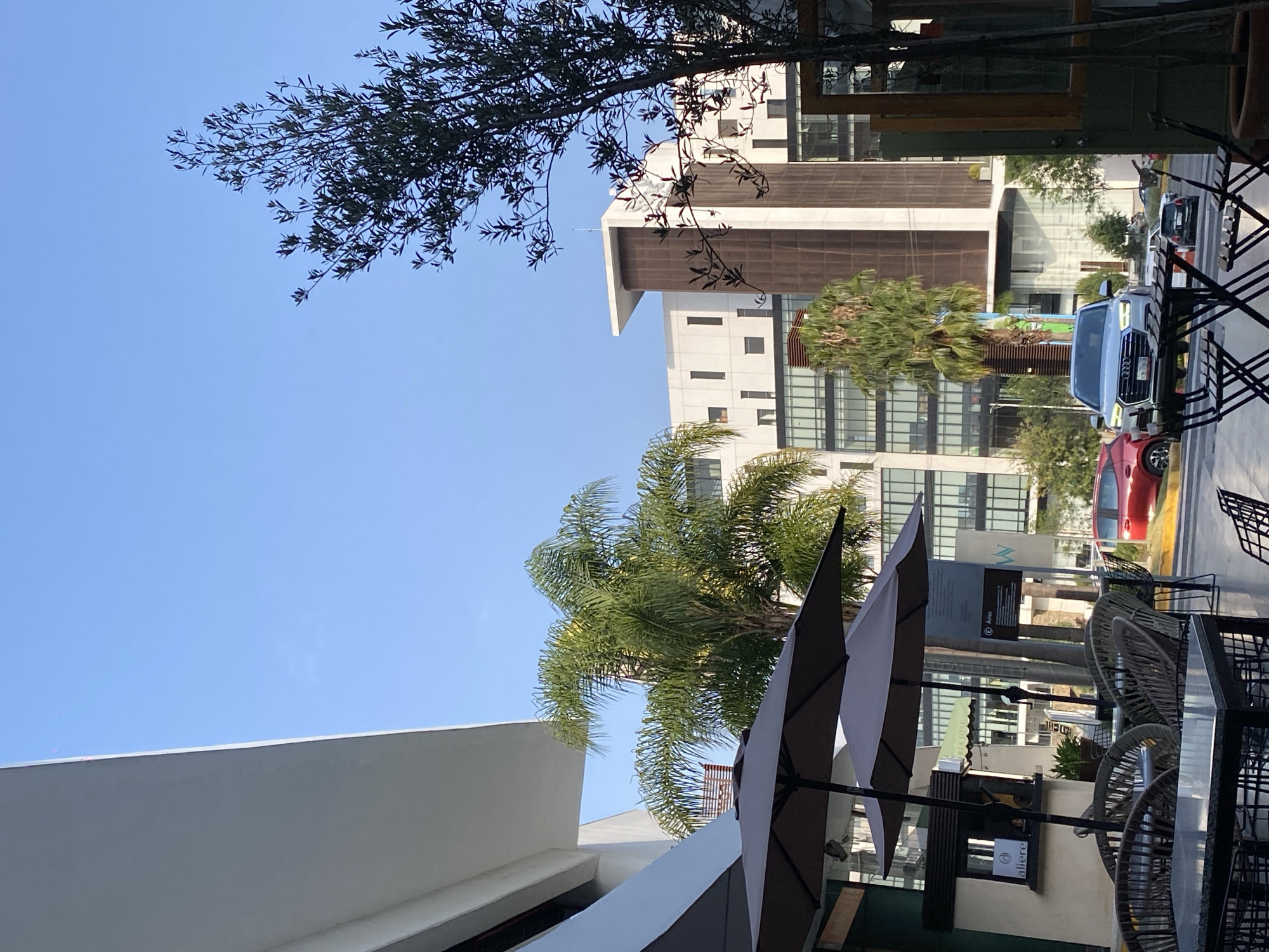 A daytime outdoor scene shows a view from a cafe or restaurant patio. The location appears to be an urban setting. In the background is a multi-story building. There are vehicles and traffic visible in the street. Nearer to the foreground are outdoor tables and chairs, along with sun umbrellas. Tree branches are visible above. The sky is blue.