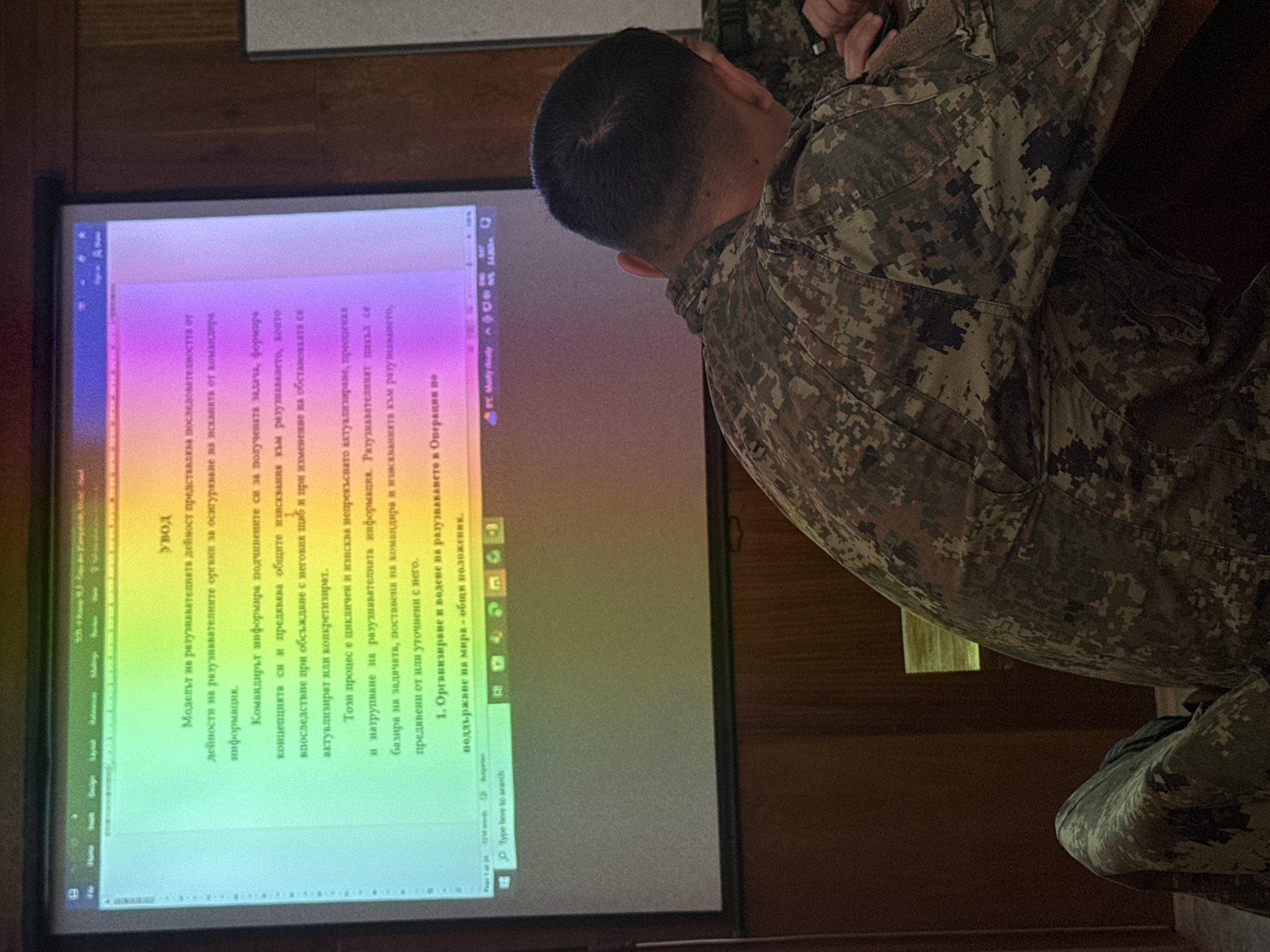 Here is a description of the image:
A man in a camouflage uniform is seen from behind, looking at a projection screen. The screen displays a document written in a Cyrillic script, likely Bulgarian. The document appears to be a presentation or a report. The overall setting appears to be a training or briefing room with wooden walls.