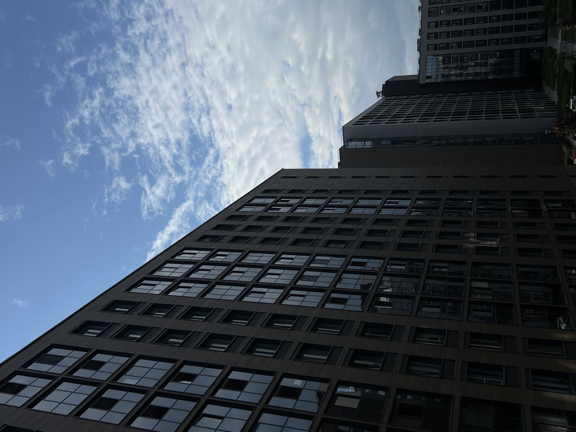 The image presents a striking upward view of modern urban architecture set against a clear, expansive sky in Saint Petersburg, Russia. The scene is dominated by two contemporary buildings, emphasizing their scale and design.
The closest and most prominent building occupies a large portion of the left and bottom of the frame. It features a dark, possibly charcoal grey or black, facade constructed with a repetitive grid pattern of numerous rectangular windows. These windows, some reflecting the bright sky and others appearing dark, create a strong visual texture. The building's diagonal orientation, angled sharply from the bottom left towards the top center, accentuates its height and modern, angular design.
Further in the upper right, a second, lighter-colored building is partially visible, appearing behind and to the side of the main structure. It also showcases a similar contemporary architectural style with multiple reflective windows. At the base of this distant building, hints of green landscaping and a partially visible light-colored car can be discerned, suggesting an adjacent street or courtyard area.
The sky is a vibrant blue, adorned with scattered white, wispy clouds that become denser and more layered towards the right side of the image, indicative of fair weather conditions. The low-angle perspective provides a dynamic composition, drawing the eye upwards along the buildings' lines into the vastness of the sky.
The scene depicts a tranquil daytime moment, likely mid-morning or afternoon, given the bright illumination and clear visibility. There are no individuals visible, and no specific activities or events are taking place, lending a sense of stillness to the urban environment. No discernible text is present within the frame.