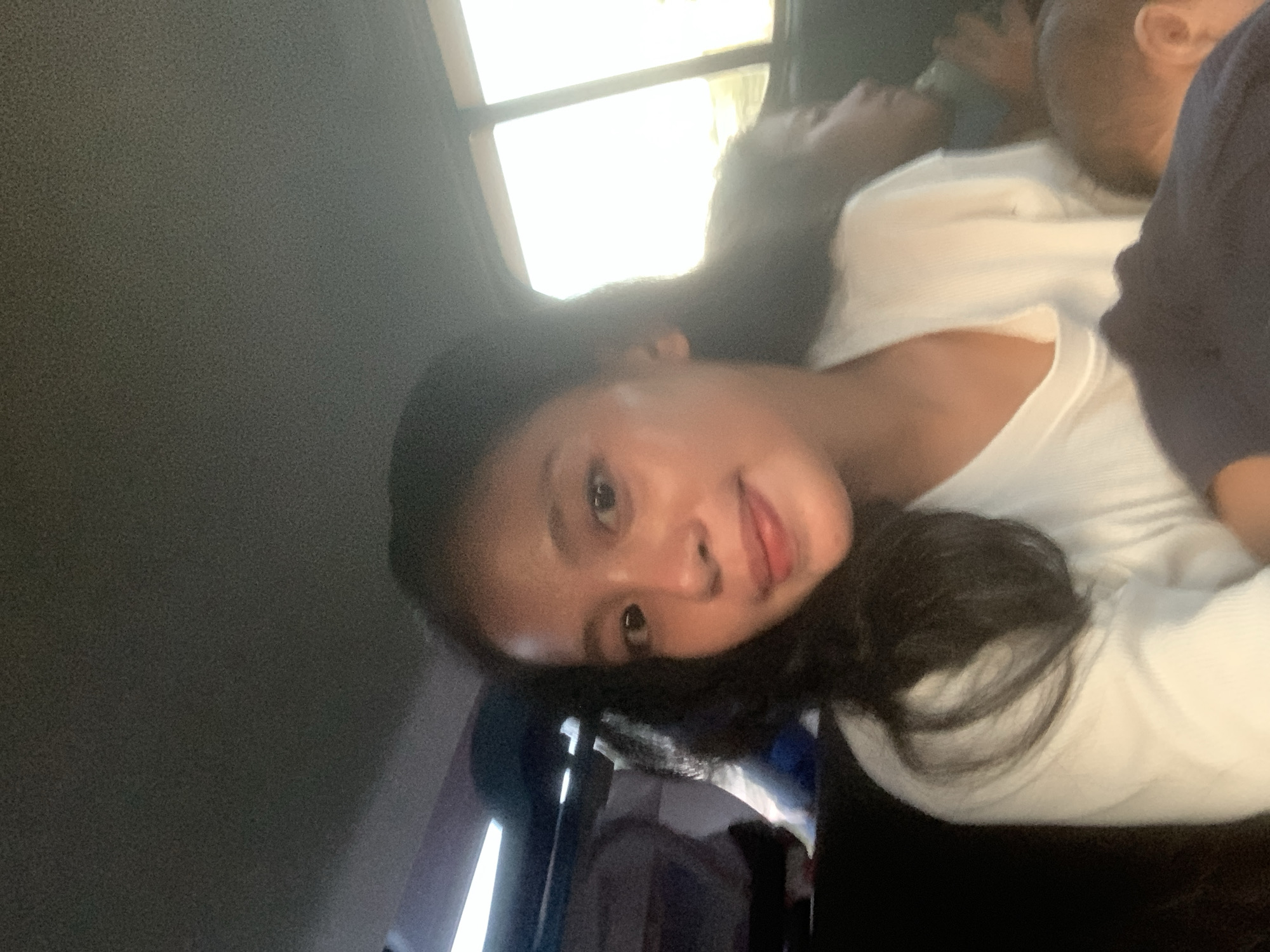 The image depicts a person with long, dark hair wearing a white shirt. There is another person visible in the background, possibly sleeping. The location seems to be indoors, likely a vehicle, based on the window visible in the background and the vehicle's ceiling. No specific indicators definitively link the scene to Batu Batu, Indonesia.