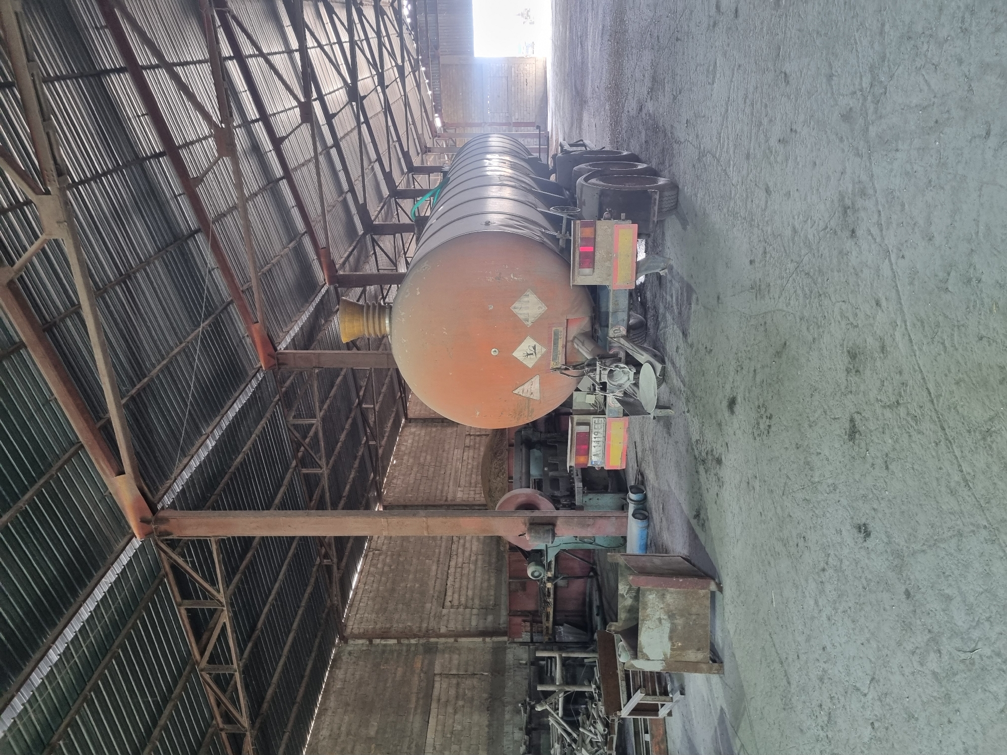 The image depicts a tanker truck parked inside a large, possibly industrial building in Ovcharovo, Bulgaria. The tanker is orange and cylindrical, resting on a truck chassis. The building has a high, corrugated metal roof supported by a metal framework. The floor is concrete. The truck appears to be stationary. The environment suggests an enclosed storage or maintenance facility.