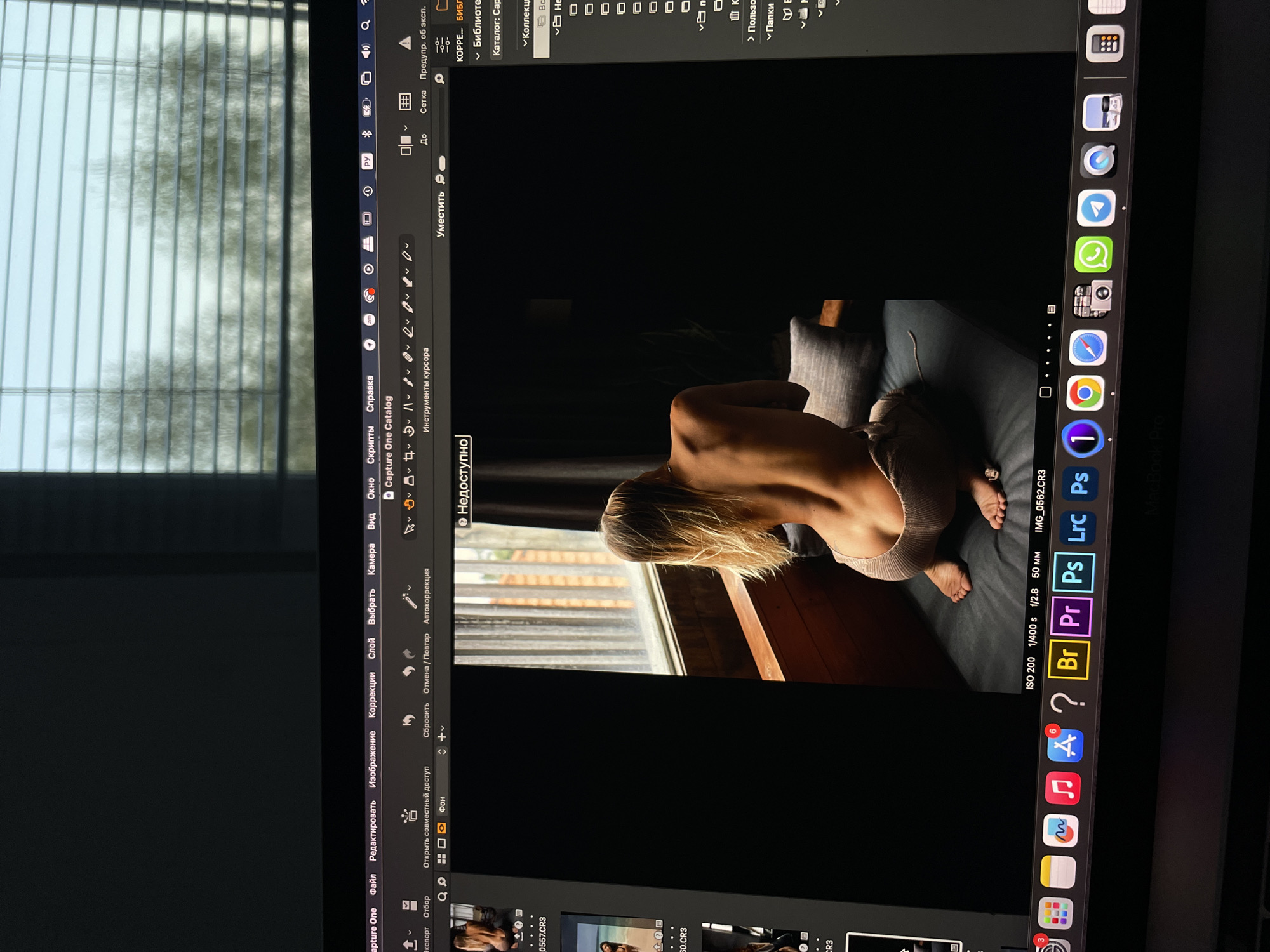 A photograph of a laptop screen showing the Capture One software with an image of a fair-skinned person from the back, leaning against a window. The background shows sunlight streaming through blinds. The laptop screen also displays the application dock at the bottom, with icons for applications such as Adobe Photoshop and Premiere Pro, along with the software's interface and tools. The laptop appears to be near a window with blinds. There is no information available in the image to suggest the city of Kazan, Russia.
