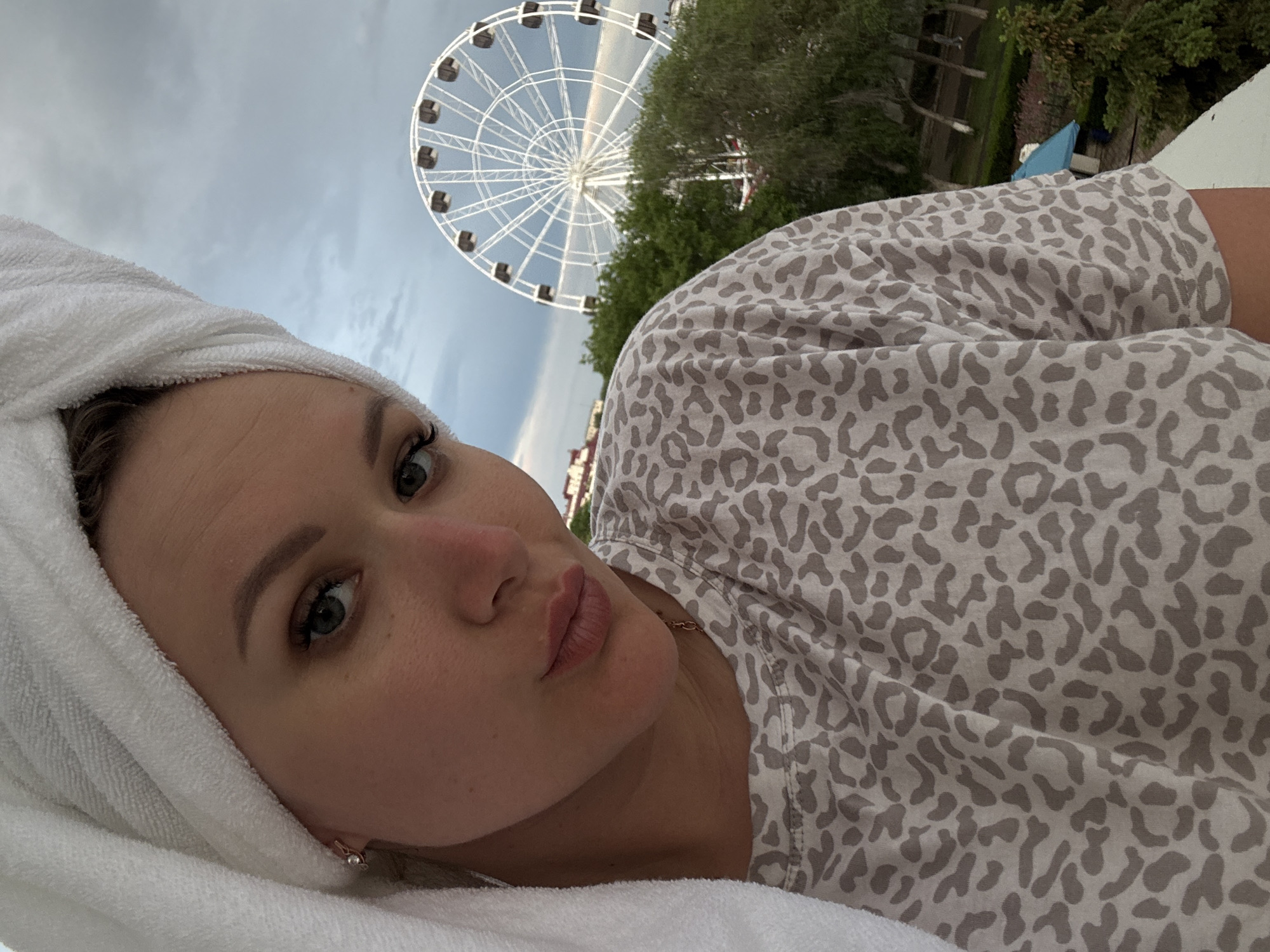 A young woman with a white towel wrapped around her head is pictured in a selfie. She is wearing a grey and white patterned shirt. Behind her, a Ferris wheel is visible, along with trees and a cloudy sky. The location is Yevpatoriya, Ukraine.