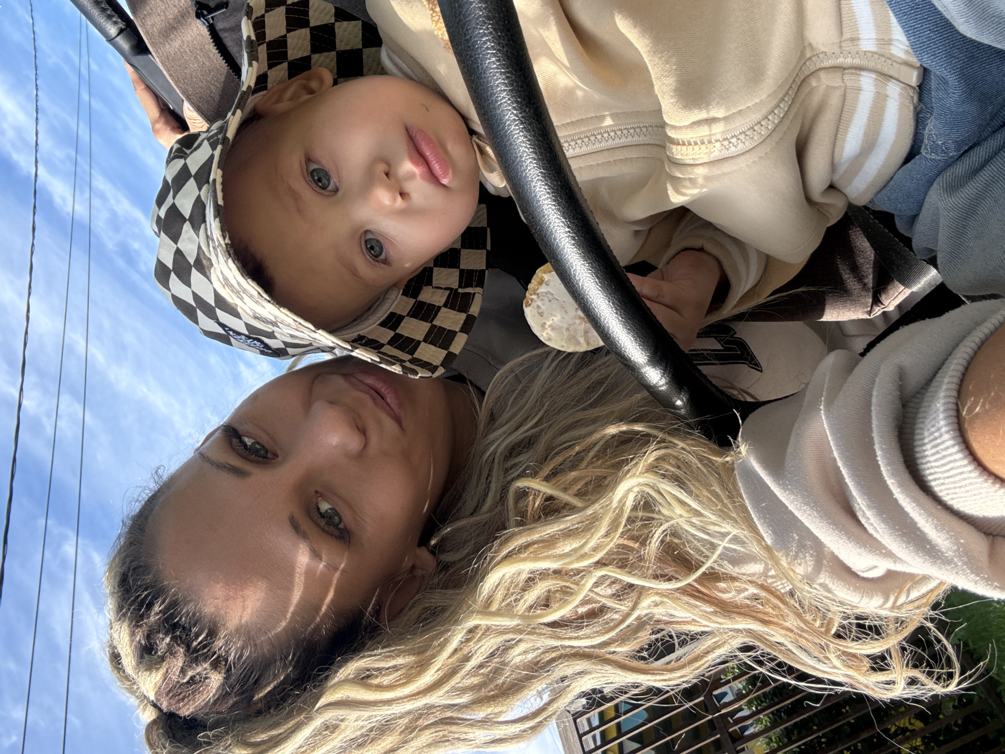 The image is a selfie taken outdoors, possibly in Zhukovo, Russia, on a sunny day with a blue sky and visible power lines. A woman with long, wavy blonde hair and striking green eyes smiles at the camera. She is wearing a light-colored hoodie. Next to her, a young child wearing a checkered bucket hat and a beige zippered jacket looks towards the camera with a slightly curious expression. The child is holding a light-colored snack. They are positioned in what appears to be a stroller, indicated by the black handlebar in the foreground. In the background, a fence and some vegetation are visible.