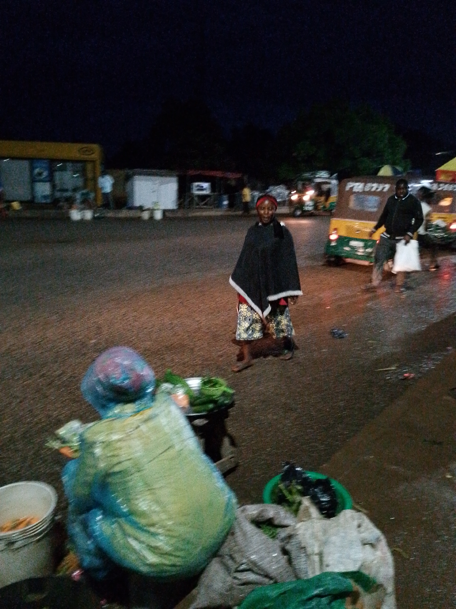 The image captures a bustling street scene at night in Bukuru, Nigeria, suggesting an active evening market or commercial area. The ground appears damp or wet, indicative of recent rain.
In the foreground, a person is hunched over, possibly a vendor, covered in a translucent blue or green plastic poncho, facing away from the camera. Beside them are various items, including white buckets, one appearing to hold orange produce (possibly carrots), and bags filled with green leafy vegetables.
In the mid-ground, a woman with a red headscarf stands facing the viewer. She wears a dark shawl over a patterned skirt and looks directly at the camera with a slight smile. To her right, a man in a dark jacket stands next to a yellow and green motorized rickshaw, commonly known as a Keke-Napep or tuk-tuk, holding a white bag. The registration number "P26777" is faintly visible on the side of the rickshaw.
The background features various dimly lit structures, including what appears to be a yellow shop or kiosk with some visible text or logos that are too blurry to decipher clearly. Other figures are scattered in the background, along with the lights of other vehicles, contributing to the overall impression of an active urban night scene. The primary activity appears to be street vending and general movement of people and vehicles.