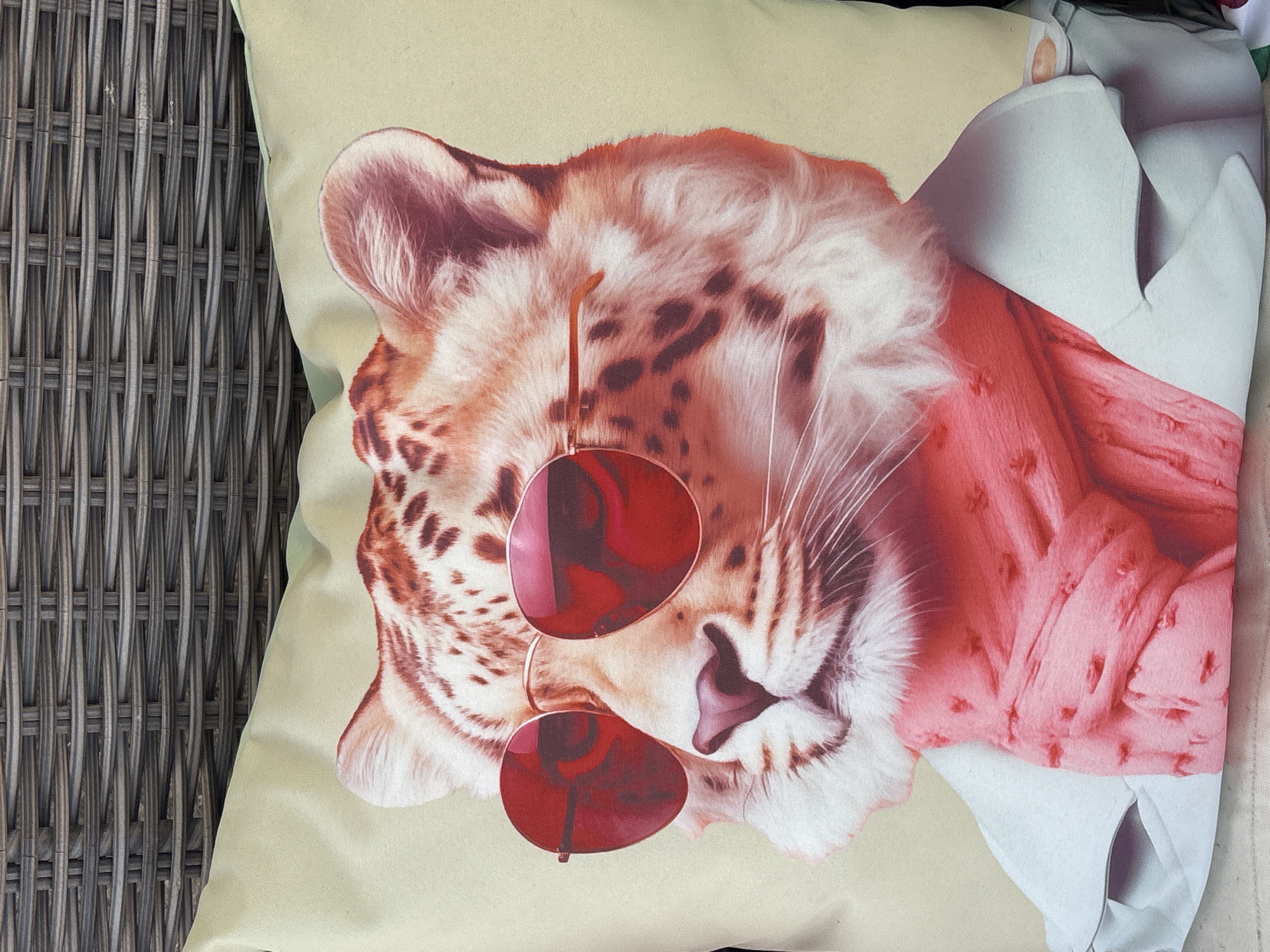 A close-up, high-angle shot shows a decorative pillow featuring a stylized portrait of a leopard wearing red-tinted sunglasses and a pink scarf. The leopard has a light-colored face with characteristic dark spots and white fur around its muzzle and ears. It is wearing a light-colored jacket over the scarf. The pillow rests on a surface that appears to be made of woven material, possibly outdoor furniture. The background is filled with the texture of this woven material, suggesting an outdoor setting. The image does not contain any elements that can be used to determine its location within the city of Orihuela, Spain.