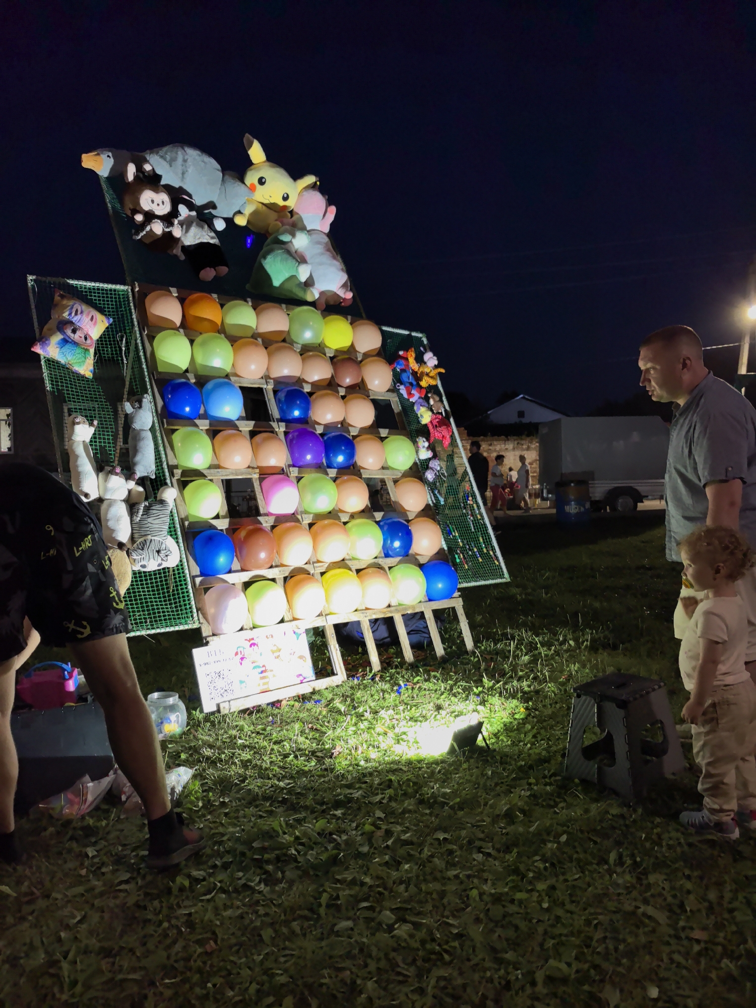 In the city of Osa, Russia, at night, a group of people are gathered around a carnival game booth. The booth features a large wooden structure with multiple rows of colorful balloons. Above the balloons, stuffed animals are displayed as prizes. A man and a young child are standing in front of the booth, facing the balloons. The man is wearing a grey t-shirt and shorts, and the child is wearing a light-colored t-shirt and pants. Another person, partially visible, is standing to the left of the booth, wearing black shorts with a white anchor design. The ground is covered in grass, and a spotlight illuminates the front of the game. In the background, there are buildings and a trailer. The sky is dark, indicating it is nighttime.