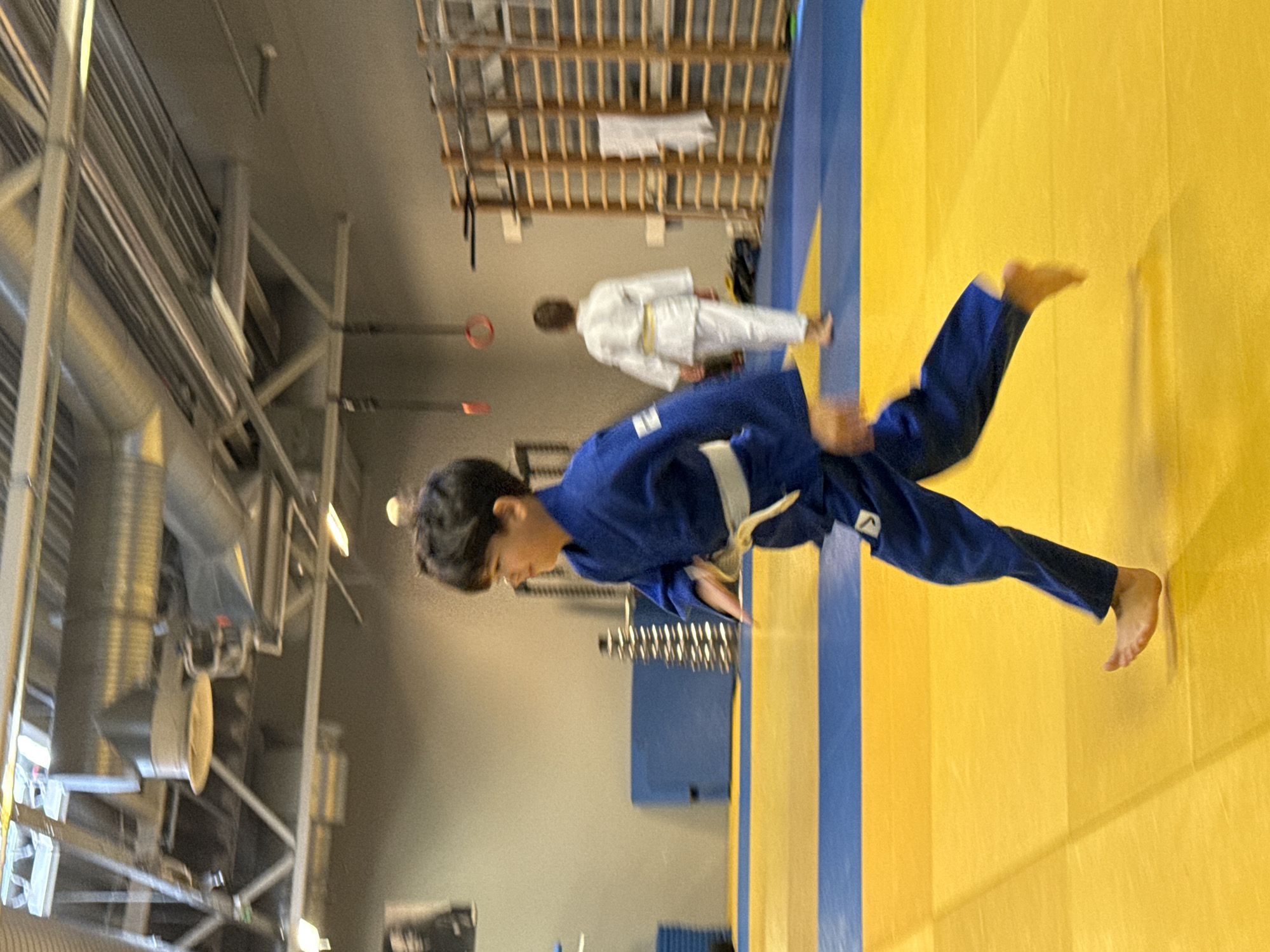 An indoor judo gymnasium is depicted. The floor is a yellow mat with blue borders. In the foreground, a young boy in a blue judogi, a traditional martial arts uniform, runs from left to right across the mat. His white belt is visible. In the background, another person in a white judogi walks away from the camera, also on the mat. The gymnasium has exposed ceiling infrastructure, including pipes and lighting fixtures. On the right wall, there is a wooden structure with horizontal bars, possibly for gymnastics or training. A framed picture is visible on the left wall. The overall lighting suggests an indoor sports facility.