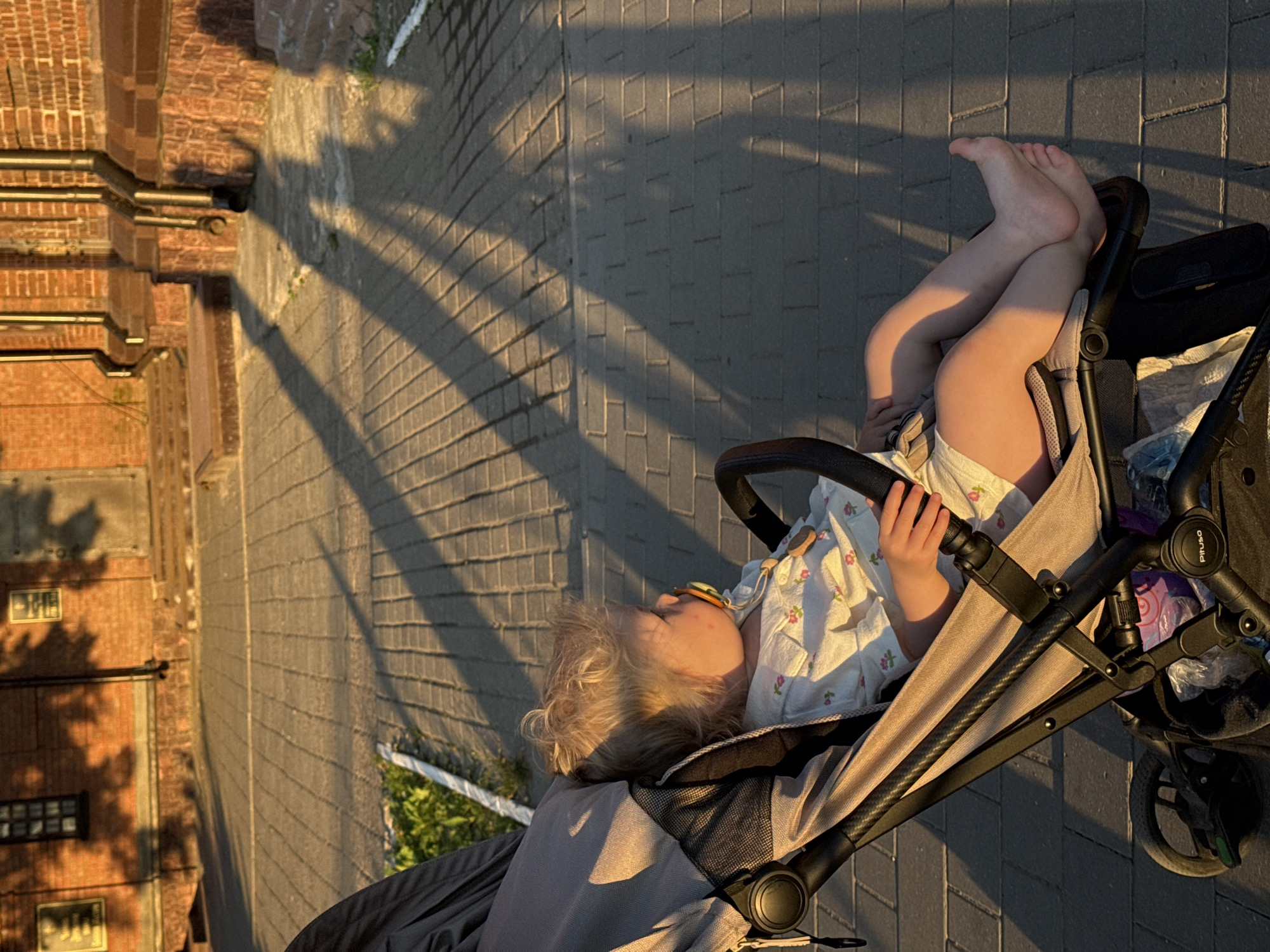 In Vladimir, Russia, at sunset, a toddler with blond hair is sitting in a stroller, facing right. The child is wearing a light-colored, floral-patterned shirt and a diaper, with a pacifier in their mouth and a wooden toy attached to a cord. The stroller is dark gray and black with a beige fabric seat. The child's legs and feet are visible, with their right leg crossed over the left. The stroller is parked on a paved walkway made of grey rectangular bricks. Long, distinct shadows of trees or architectural elements are cast across the walkway, indicating late afternoon or early evening. In the background, a red brick building with a window and architectural details is visible, illuminated by the warm light of the setting sun. The overall scene suggests a peaceful moment outdoors.
