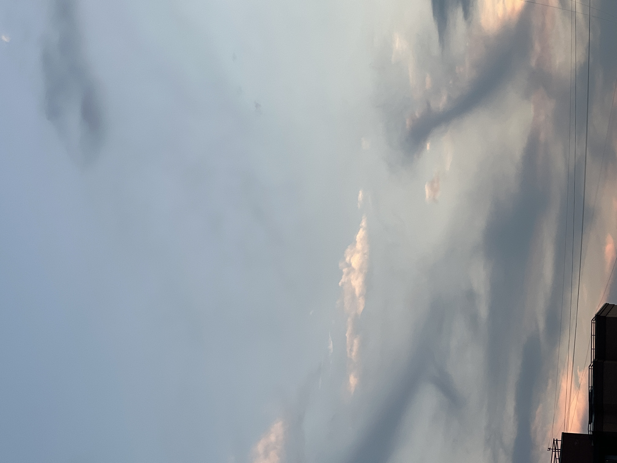 The image is a wide shot of a twilight sky over a city. The sky transitions from a pale blue at the top to a lighter, softer blue towards the horizon, with scattered clouds in various shades of gray and white. Some of the clouds have a pinkish hue, suggesting the sun is setting or has recently set. In the lower portion of the image, the rooftops of buildings are visible, with some dark gray clouds obscuring the upper parts of the structures. Thin electrical wires are stretched horizontally across the lower part of the frame. The overall impression is a serene, urban evening scene.