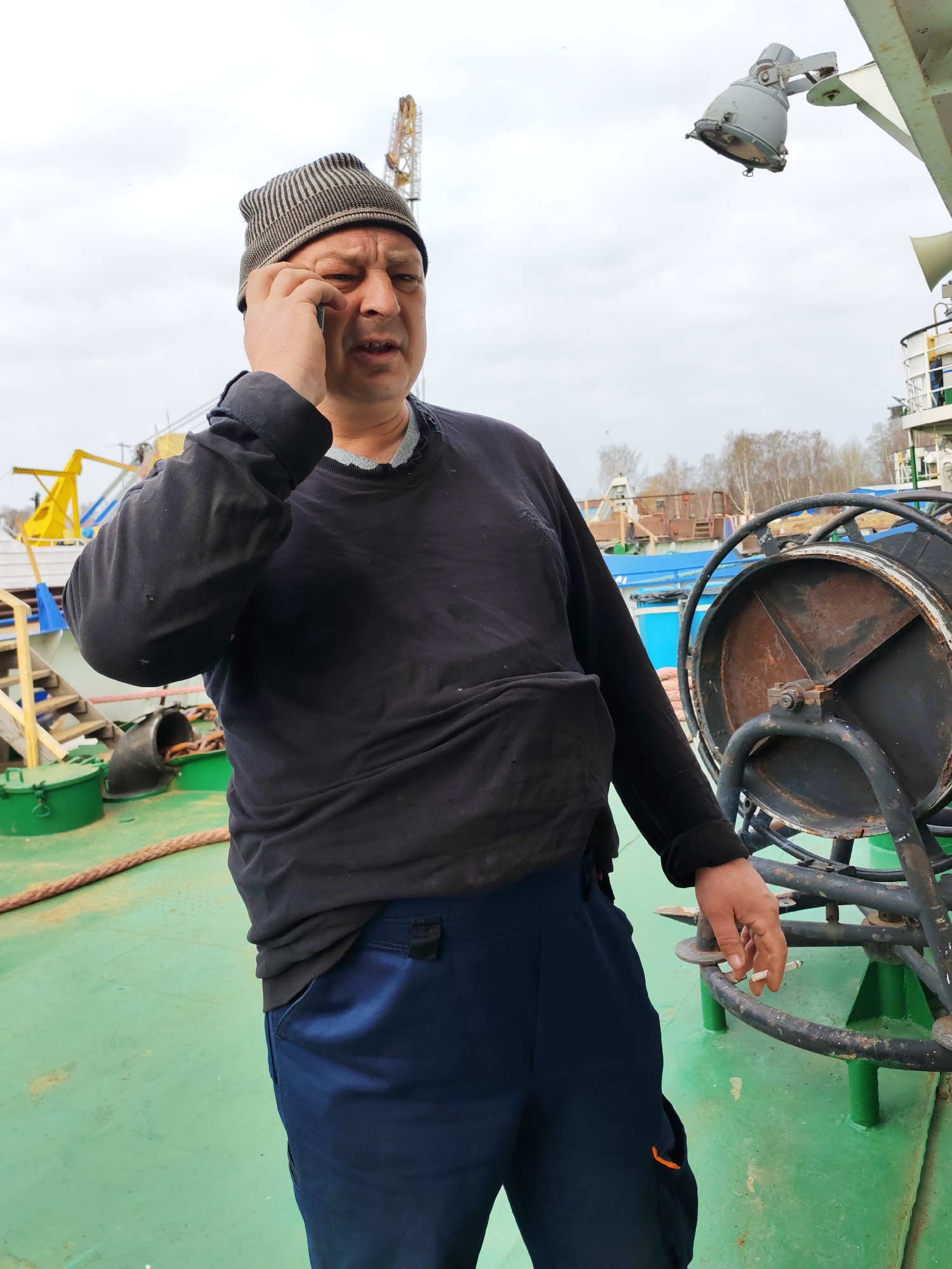 A middle-aged man wearing a striped beanie, a black long-sleeve shirt, and blue pants stands on a green deck with industrial equipment, possibly on a boat or ship. He is holding a cigarette in his right hand and has his left hand up to his ear as if using a phone. The man appears to be outdoors under an overcast sky. In the background are trees, a light-colored sky, and hints of industrial infrastructure. Given the prompt, the location is likely Rybinsk, Russia.