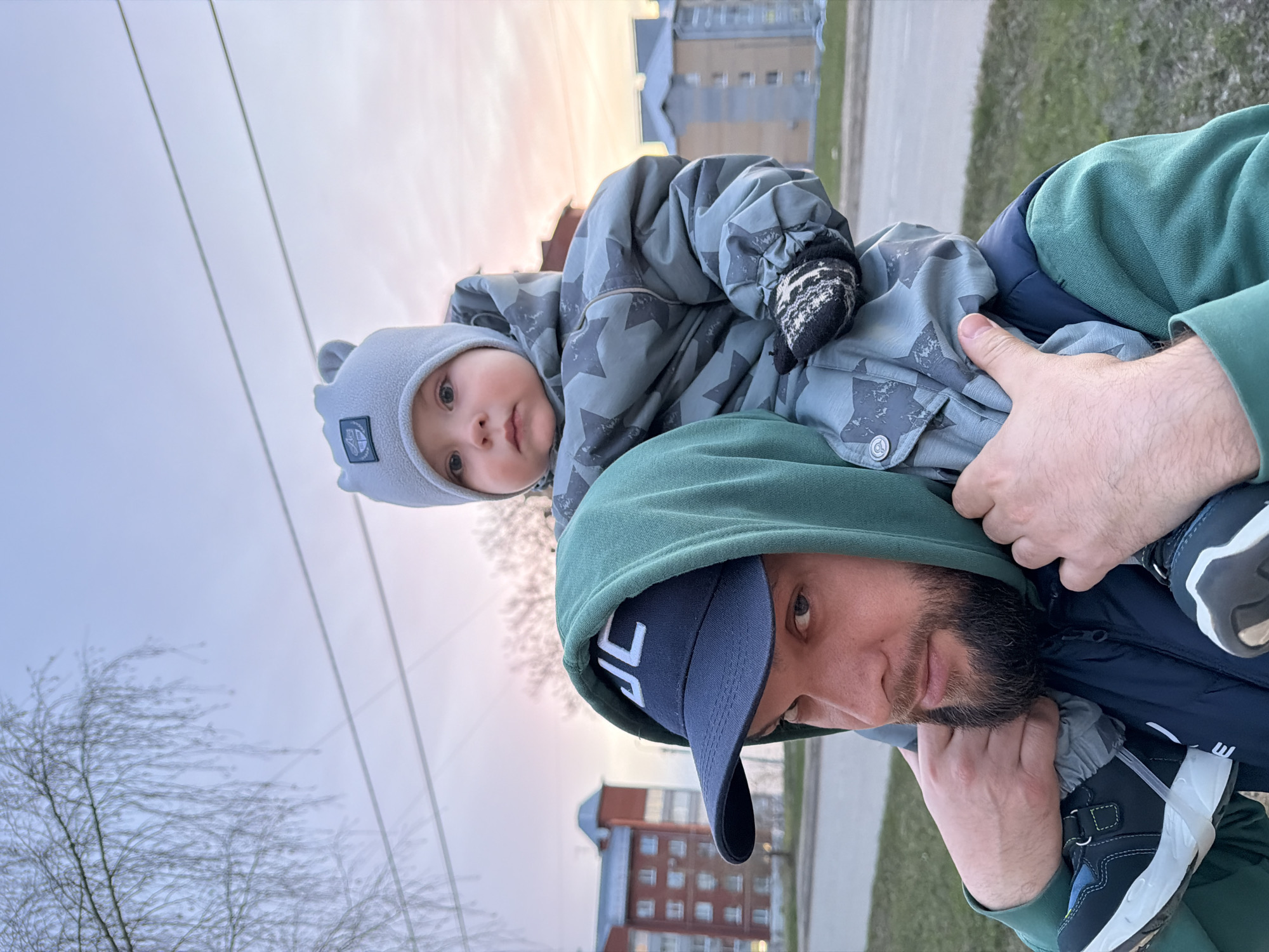 The photo depicts a man carrying a young child on his shoulders, likely in Votkinsk, Russia, based on recognizable architectural styles. The man, wearing a green hooded sweatshirt and a blue baseball cap, looks directly at the camera. He has a beard and appears to be holding the child's feet or legs with his hands. The child, wearing a gray hat and a patterned gray snowsuit with mittens, sits on the man's shoulders and also looks towards the camera. A multi-story residential building is visible in the background. The sky appears overcast, and bare trees are in the foreground on the left. A paved walkway is visible, running diagonally from the background to the lower right. The lighting suggests either early morning or late afternoon.
