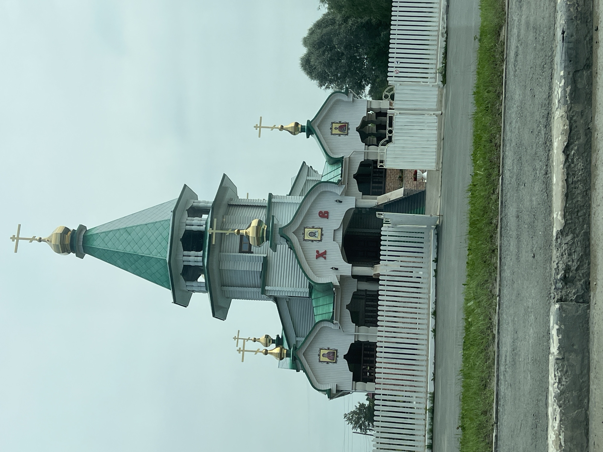 The image shows an Orthodox church with a green roof and a golden dome, located in the city of Troitskoye, Russia. The church is made of white wood and features multiple crosses on its rooftops. It is surrounded by a white picket fence. In front of the church is a patch of green grass, and a road is visible in the foreground. The sky is overcast. No people or any discernible actions are depicted in the image.