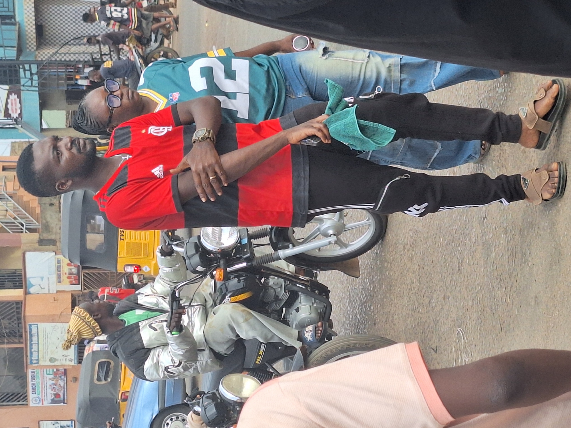 In Sabon Tasha, Nigeria, a man wearing a red and black Adidas shirt and black Adidas track pants with white stripes is standing on the sidewalk next to a motorcycle. He is wearing brown sandals and a gold wristwatch and has a green cloth in his hand. Another person in a green football jersey is leaning against him. A motorcyclist with a yellow and tan striped hat is seated on the bike. In the background are buildings, a yellow TVS three-wheeled vehicle, and other pedestrians.