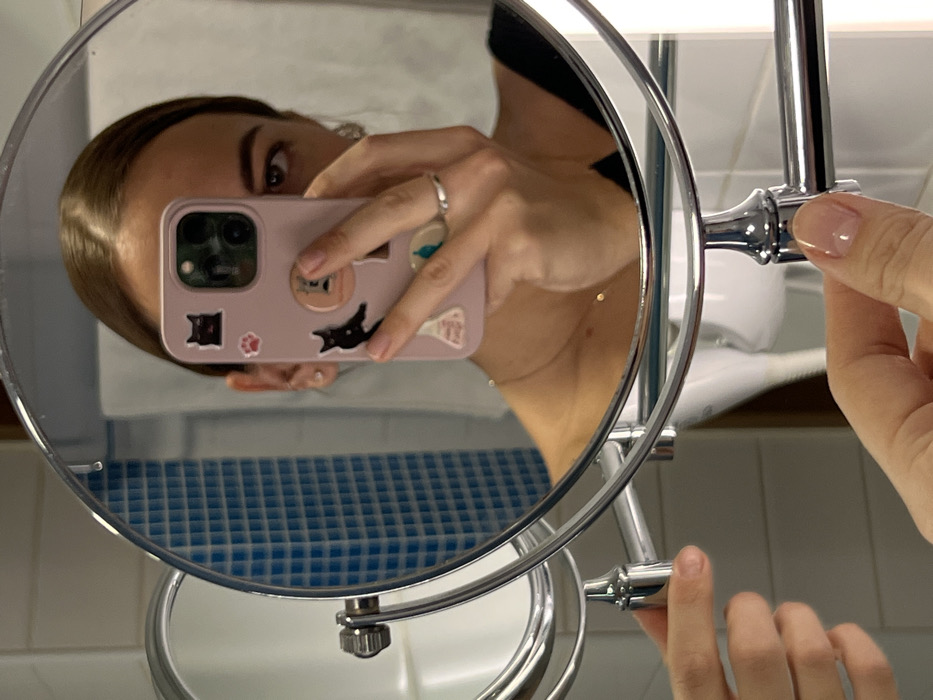 A person is reflected in a chrome, circular makeup mirror. The reflection includes the individual's face, shoulder, and hand holding a pink cell phone. The phone case is decorated with cat stickers. A silver ring is visible on one of the person's fingers. Another hand adjusts the mirror’s position. The background features a tiled wall and a white countertop or sink.