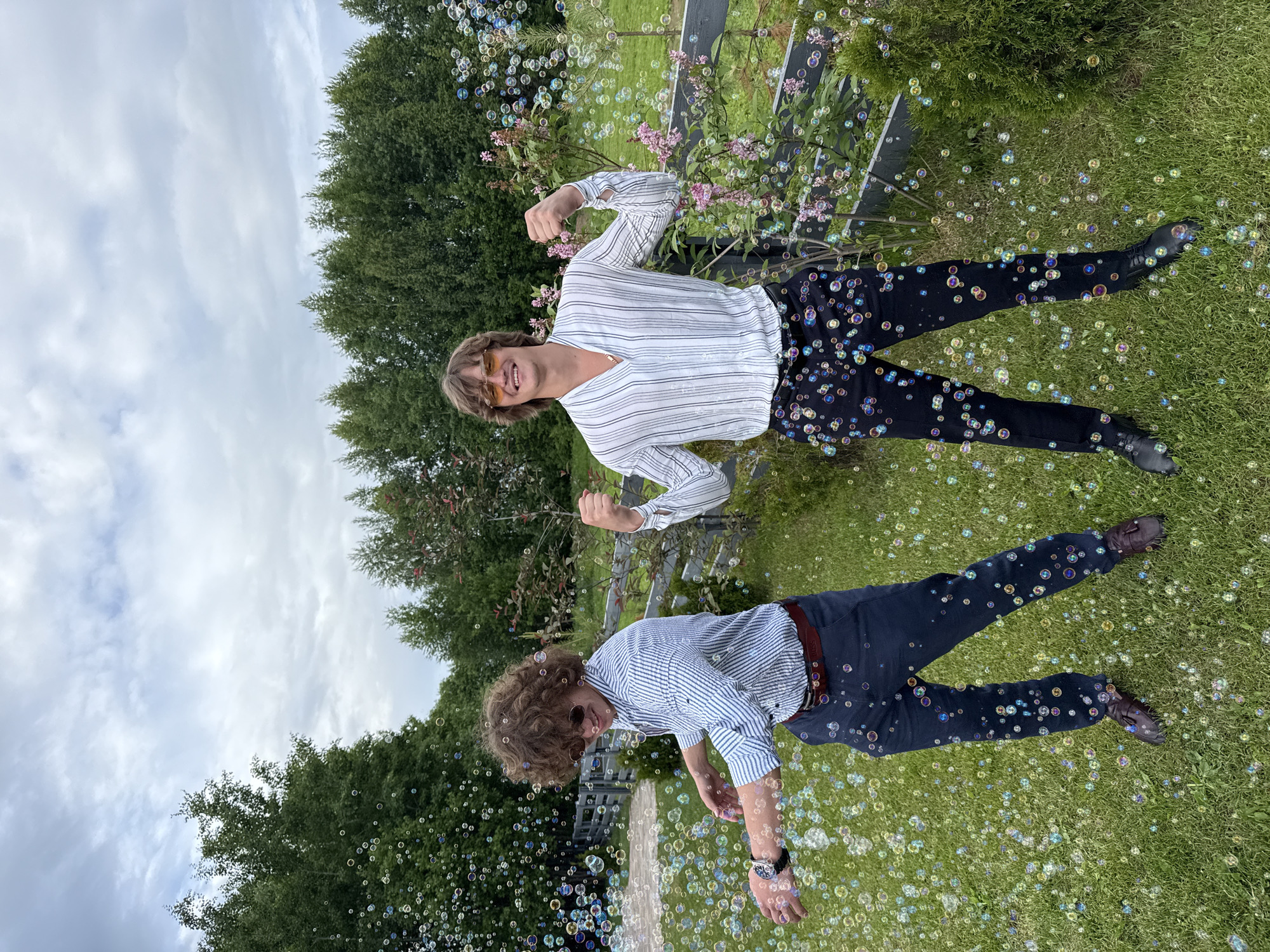 Two young adult males are depicted outdoors in the city of Perm, Russia, amidst numerous iridescent soap bubbles floating across a green lawn, against a backdrop of trees and an overcast sky. The subject on the left, with curly brown hair and sunglasses, wears a light blue and white striped long-sleeve shirt, dark blue trousers with a brown belt, and brown tassel loafers, with a watch on his left wrist. He is captured mid-action, smiling downwards, with his arms extended as if interacting with the bubbles. The subject on the right, with medium-length light brown hair and yellow-tinted sunglasses, wears a white long-sleeve shirt with thin black vertical stripes and a V-neck, dark trousers, and black shoes, with a gold chain visible. He smiles broadly at the viewer while flexing both biceps. Behind the individuals, a dark gray wooden fence separates the manicured lawn from a line of dense green trees, including both deciduous and evergreen varieties. Flowering plants with pink blossoms are visible to the right of the fence. A light-colored paved area is partially visible on the far left. The sky is uniformly cloudy, indicating an overcast daytime setting with diffused lighting.