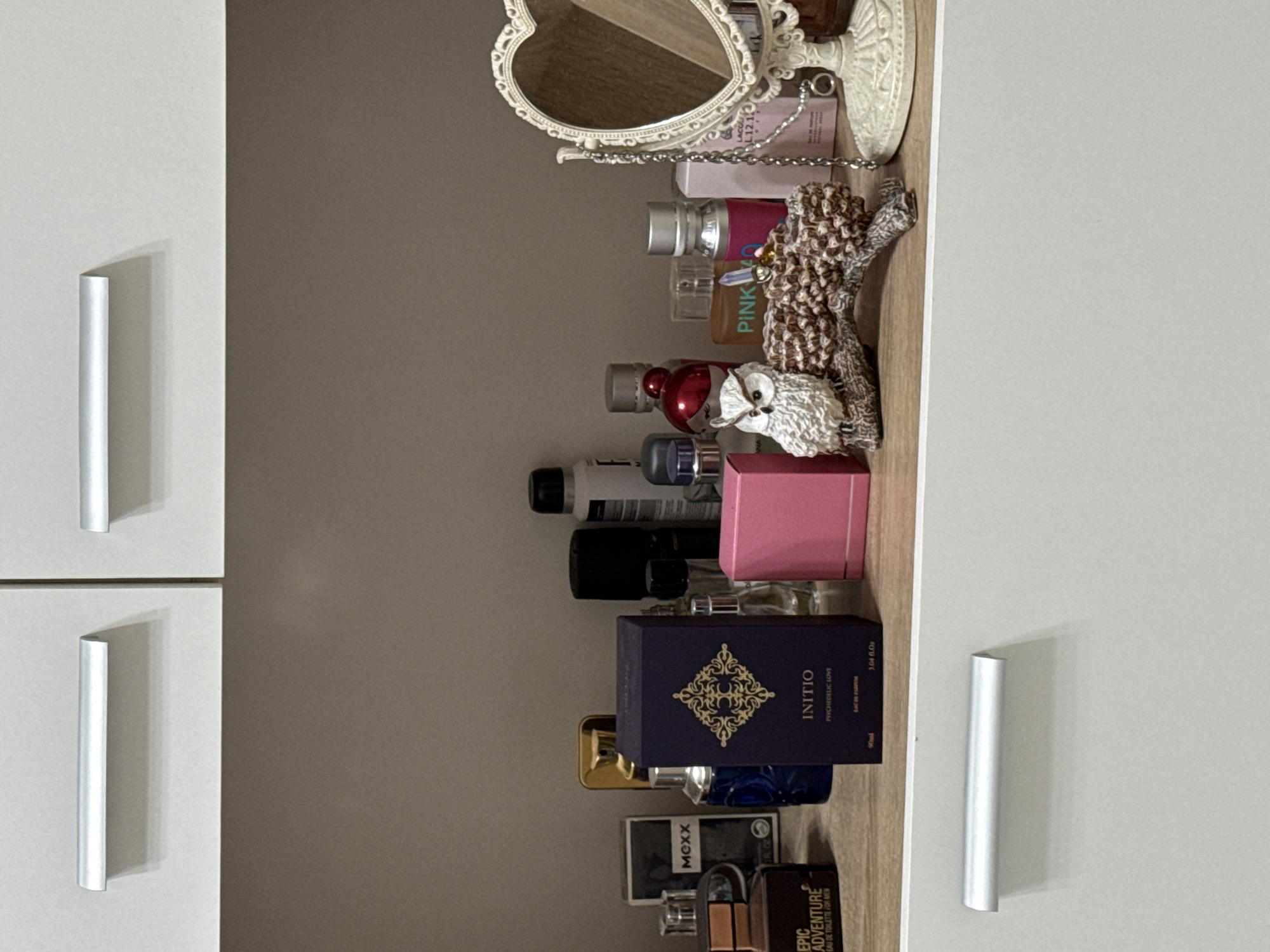 This image shows a shelf with various items on it. Among these items are a mirror, bottles of perfume, and a small figurine of an owl perched on a decorative object. The background suggests the shelf is part of a cabinet or storage unit. The style of the objects and the storage unit indicates the scene is in a domestic environment. The location in the city of Unknown, Unknown cannot be determined.