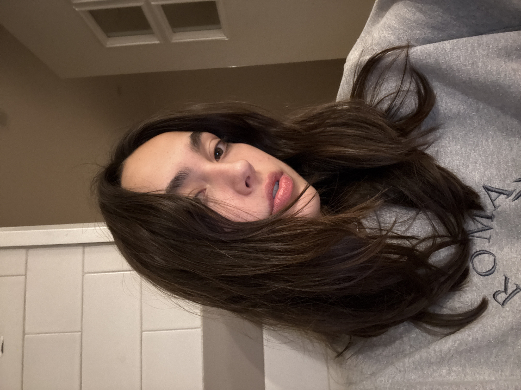 A young woman with brown hair is wearing a gray sweatshirt and lying down. She has fair skin, brown eyes, and full lips. The background consists of a tiled wall and ceiling, with a window grid visible in the top left corner. It is difficult to determine if the location is Mexico City, Mexico, based on the given information.