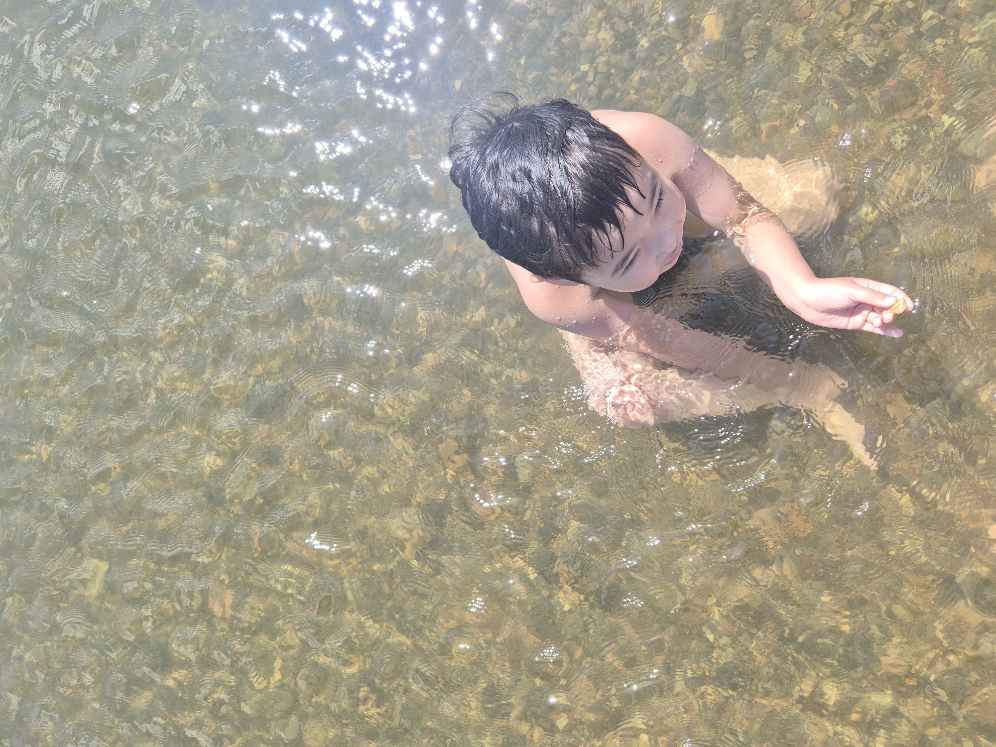 A young boy with dark hair is standing in shallow, clear water with a rocky bottom. The sun is shining brightly, creating reflections on the water's surface. The boy is looking down at his hand, which is holding a small, light-colored stone. He is wearing swim trunks, and only his upper body and arms are visible. The scene appears to be in a natural body of water, possibly a river or stream, in the city of Novoishimsky rural district, Kazakhstan.