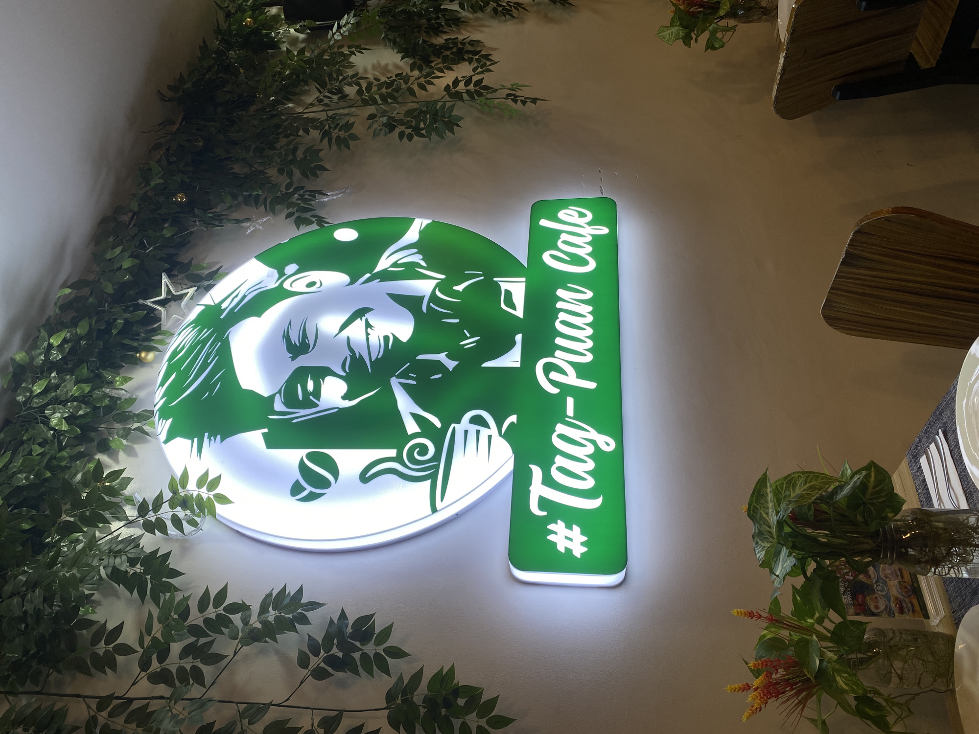 The image depicts the interior of a cafe, likely "Tag-Puan Cafe" in Davao City, Philippines, based on the signage. A prominent, circular green neon sign features a stylized portrait of a smiling man, a coffee cup, and coffee beans, with the text "#Tag-Puan Cafe" displayed below it in white neon letters on a green rectangular sign. Green artificial foliage hangs from the ceiling and walls, creating a lush atmosphere. In the foreground, a table is set with placemats, cutlery, plates, and two glass vases containing plants and floral arrangements. Wooden chairs are visible in the background. The lighting is warm and ambient, typical of a dining establishment.