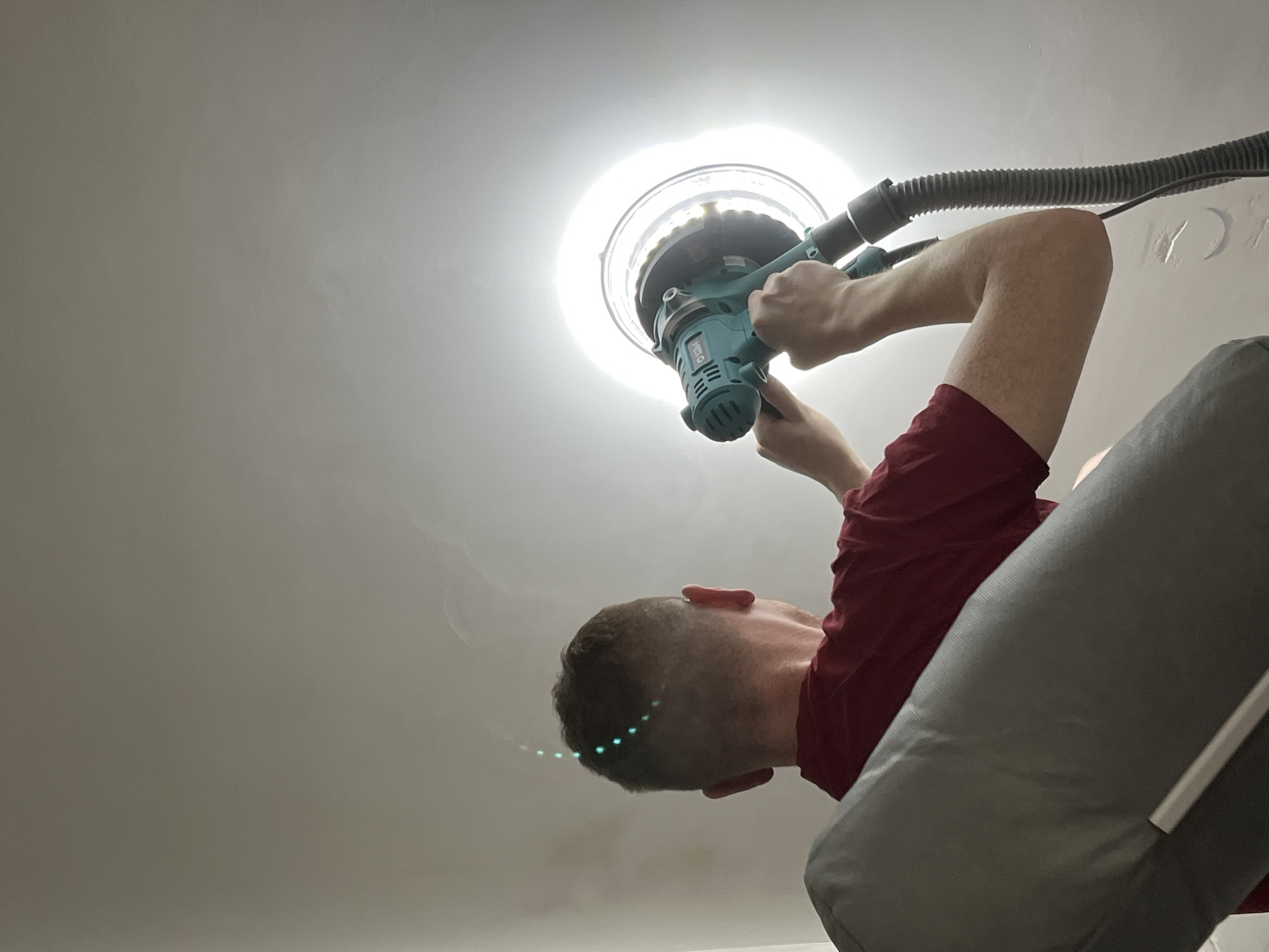 A man in a red t-shirt is sanding a ceiling with a power sander. The sander is teal and has a large circular sanding head with a vacuum hose attached. The man is standing on a stepladder. The ceiling is white and appears to have been recently plastered. The location is likely an interior space under renovation in Verkhnyaya Pyshma, Russia, although there is no visual confirmation of this.