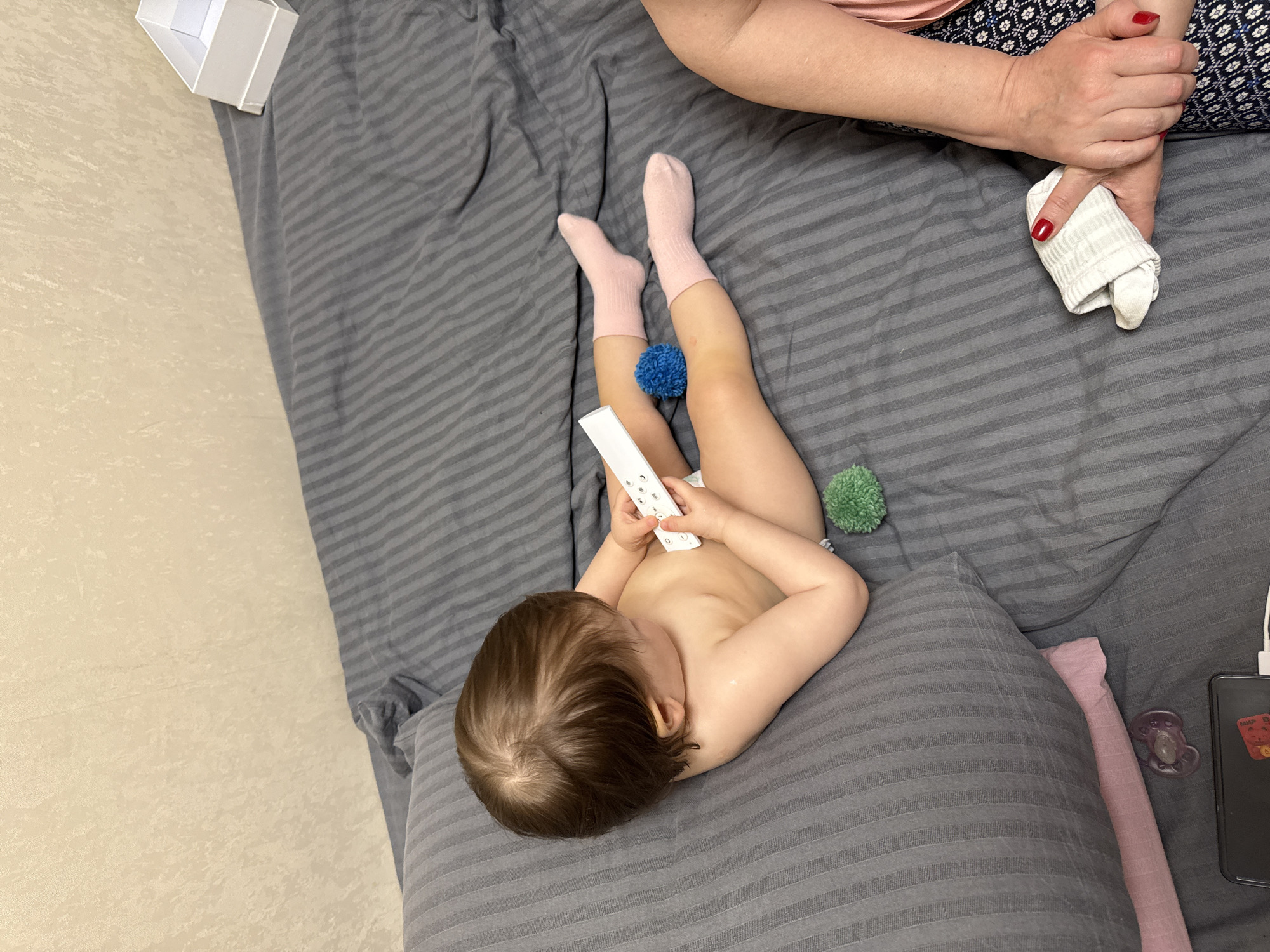 This overhead shot captures an intimate indoor scene featuring a toddler and an adult. The toddler is lying on their back on a grey, striped comforter, their head resting on a matching pillow. They have light brown hair and are wearing pink socks, with their torso bare and a diaper visible. In their hands, the toddler holds a white remote control. A blue and a green fluffy pom-pom are placed on the bedding near the toddler's legs.
Partially visible in the upper right is an adult, whose arms and hands are in view. The adult wears a patterned garment and has red nail polish, holding a rolled-up white cloth or sock. To the left, a light-colored floor or wall is visible, with a small open white box situated on the edge of the bedding.
Near the adult's side, a dark smartphone is seen, connected to a white charging cable. Adjacent to the phone are a purple pacifier and a pink fabric item. A sticker or card on the back of the smartphone displays the partially visible text "МИР". The scene conveys a relaxed, domestic atmosphere, likely a casual moment between a child and a caregiver during the day.