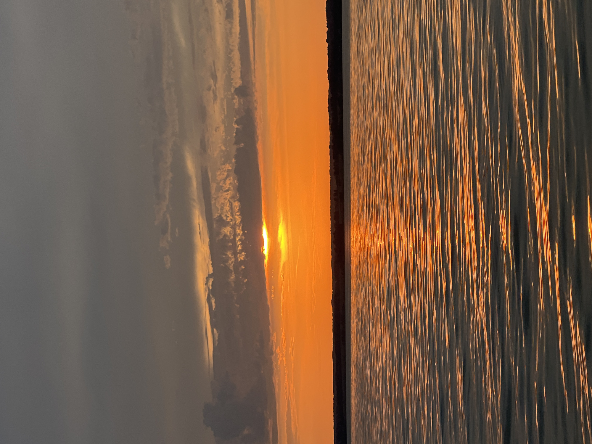The image depicts a sunset over a body of water, likely a lake or sea, near the city of Beregovoe, Russia. The sky is a vibrant mix of orange and gray, with scattered clouds. The sun, partially obscured by clouds, casts a warm glow across the horizon. The water's surface is rippled, reflecting the colors of the sky and creating a shimmering effect. A dark, silhouetted treeline is visible on the distant shore. There are no people or specific objects of interest in the foreground. The overall scene is tranquil and picturesque, characteristic of a sunset in a natural setting.