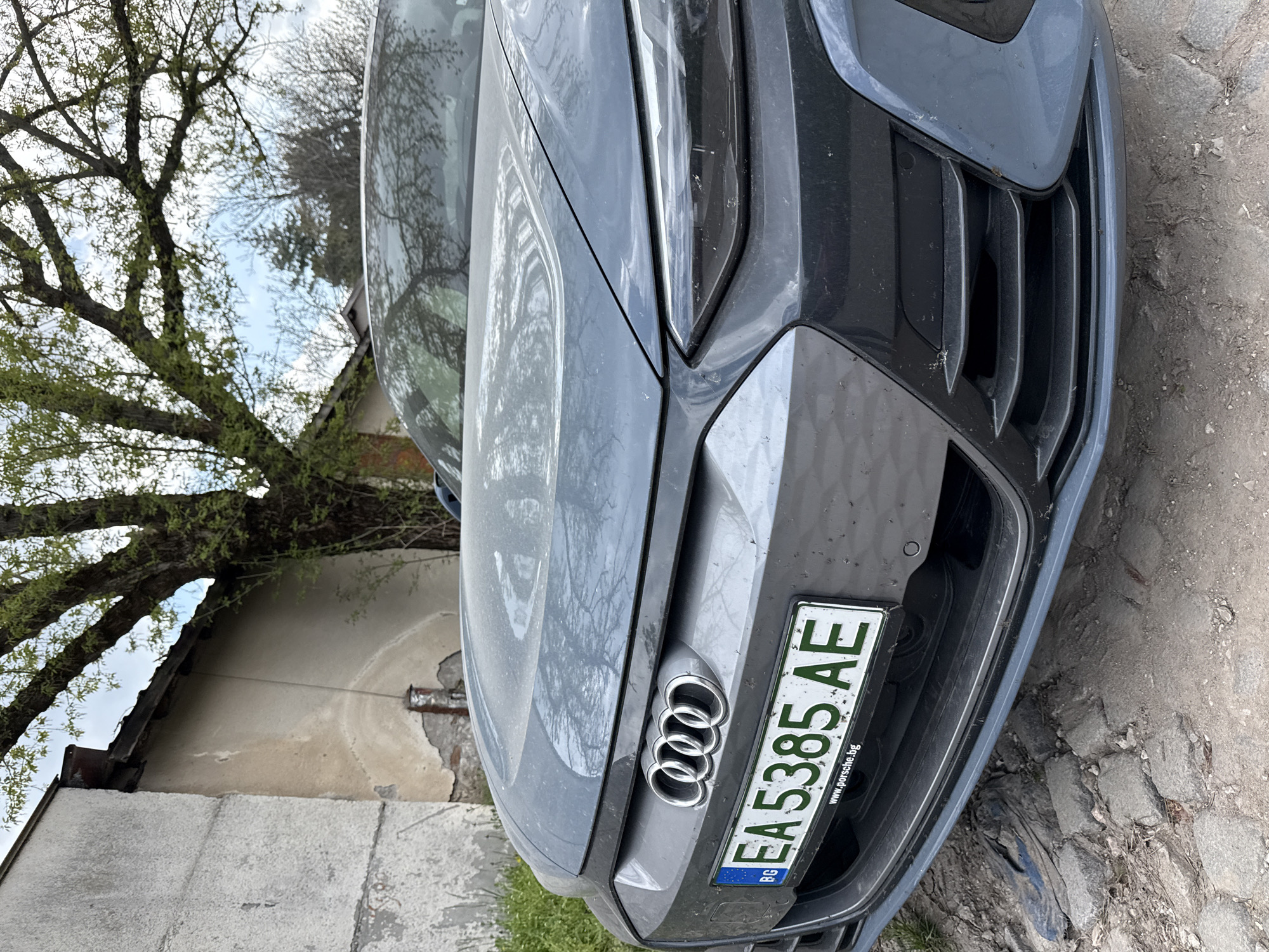The image shows the front of a gray Audi car parked on a cobblestone surface. The car has a Bulgarian license plate "EA 5385 AE". In the background, there is a tree with green foliage and a building with a light-colored exterior. The website "www.porsche.bg" is visible on the license plate frame. The location is Haskovo, Bulgaria.